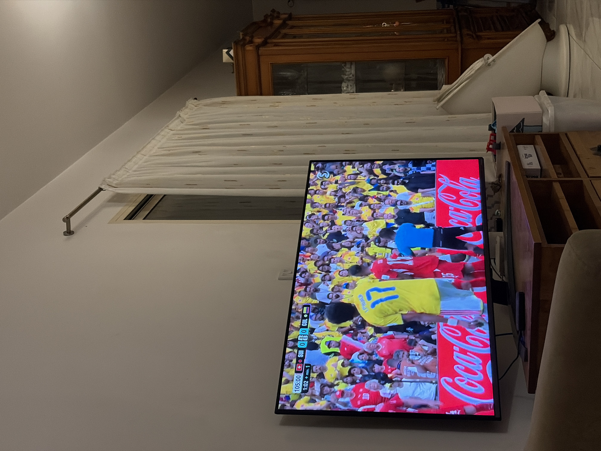 In a dimly lit room, a large flat-screen television displays a live broadcast of a soccer match. The game's score is visible on the screen: 1:02 + min. 1, with Switzerland (SUI) at 0 and Colombia (COL) at 0. The players on the screen are wearing yellow and red jerseys. In the foreground, a wooden entertainment center supports the television. To the right of the television, a tall, wooden cabinet with glass doors stands against the wall, and a white curtain hangs beside it. The overall environment suggests a domestic setting, likely a living room, in Germasogeia Municipality, Cyprus.
