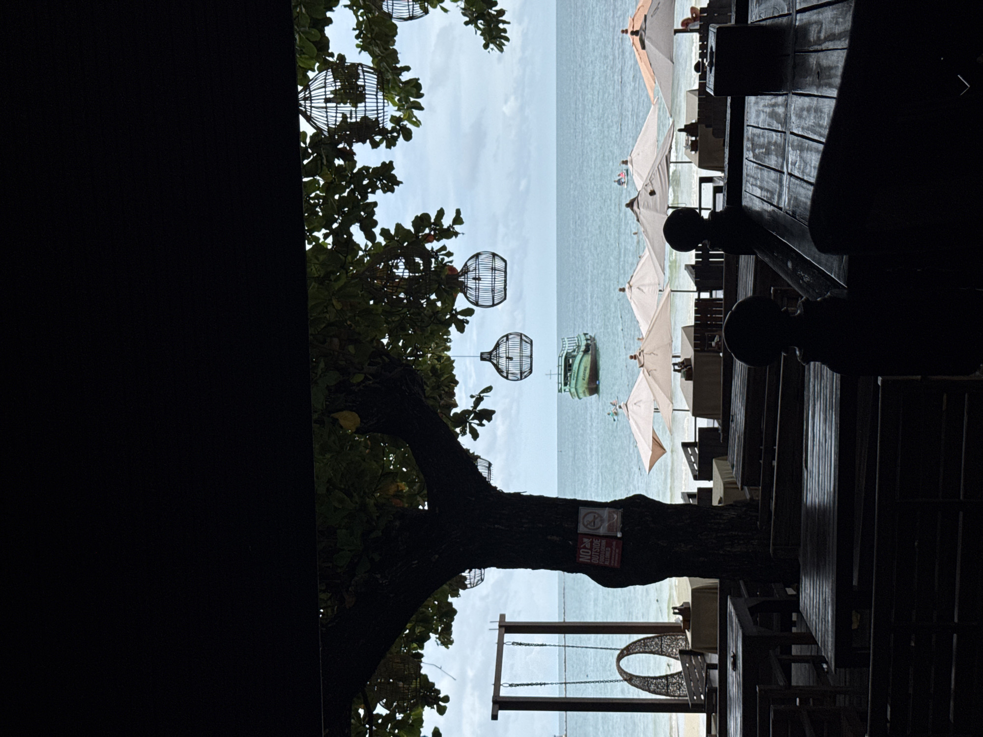 The image depicts a view from a covered outdoor seating area looking out towards Ao Phai beach in Ko Samet, Thailand. The foreground is dominated by dark silhouettes of wooden tables and benches, suggesting a restaurant or cafe. A large tree with dark foliage stands prominently to the left, its branches adorned with decorative cage-like lanterns. To the left of the tree, a wooden swing set with a woven hanging seat is visible. A sign attached to the tree trunk reads "NO OUTSIDE FOOD OR DRINK ALLOWED".
Beyond the seating area, the sandy beach is lined with cream-colored umbrellas and tables. The ocean stretches out to the horizon under a cloudy sky. A green boat is anchored in the water, and two small figures can be seen on a paddleboard in the distance. The overall scene conveys a relaxed, tropical atmosphere with a hint of the day's activities on the beach.
