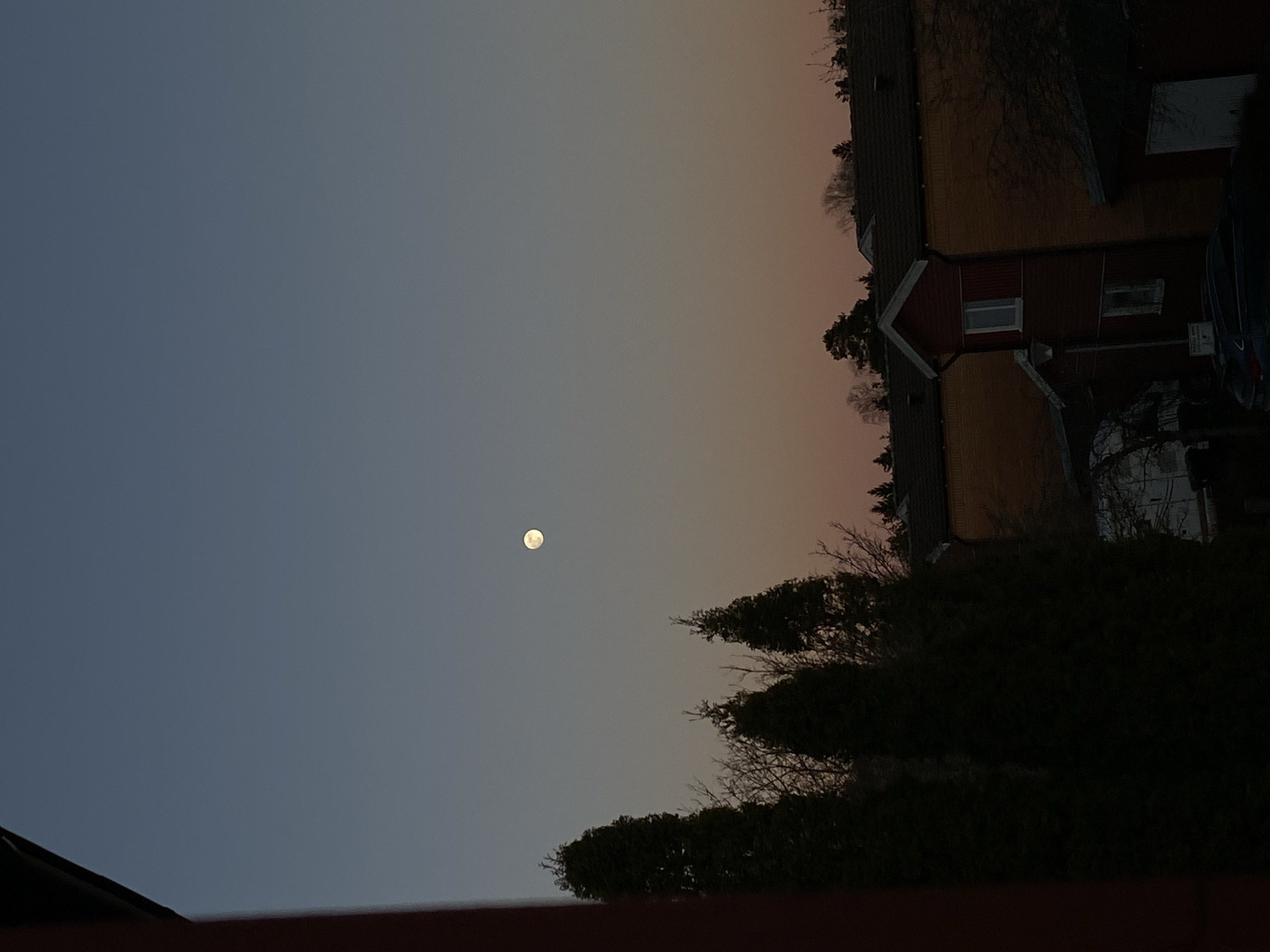 The image shows a nighttime scene in Björklinge, Sweden. A full moon is visible in the sky, which transitions from a dark blue near the bottom to a light orange-pink near the top. The horizon line features silhouettes of trees and the corner of a red wooden house with a roof and what looks to be a carport in the foreground. A car can be seen in the driveway. The overall lighting is low, with the moon being the primary light source. The picture is taken from a low angle.