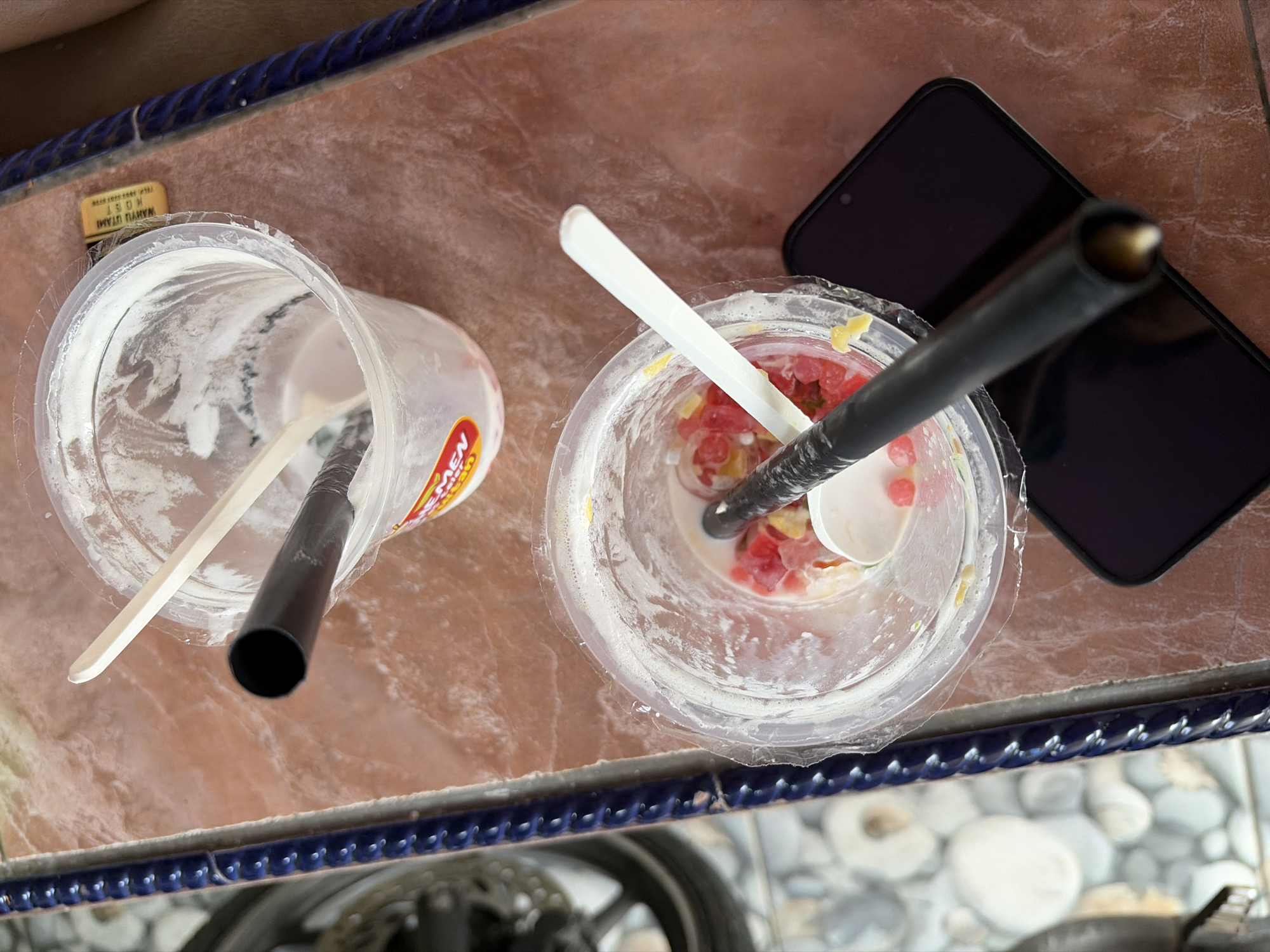 The image depicts two empty plastic cups, likely containing remnants of a sweet treat such as yogurt or ice cream, on a reddish-brown tiled surface. Both cups have used wooden spoons inside. One cup has a black plastic straw resting on its rim, partially obscuring the cup's contents, which appear to include small pieces of red fruit. The second cup, positioned above and to the right of the first, also has a straw and a wooden spoon. The background shows a blurred motorcycle wheel and a textured blue metal railing. A dark smartphone is visible beneath the lower cup, suggesting it was used as a surface. The scene is likely outdoors, given the presence of a motorcycle and the overall lighting. Based on the information provided about the location being Boja, Indonesia, the environment suggests a casual setting, possibly a street vendor or an outdoor cafe.