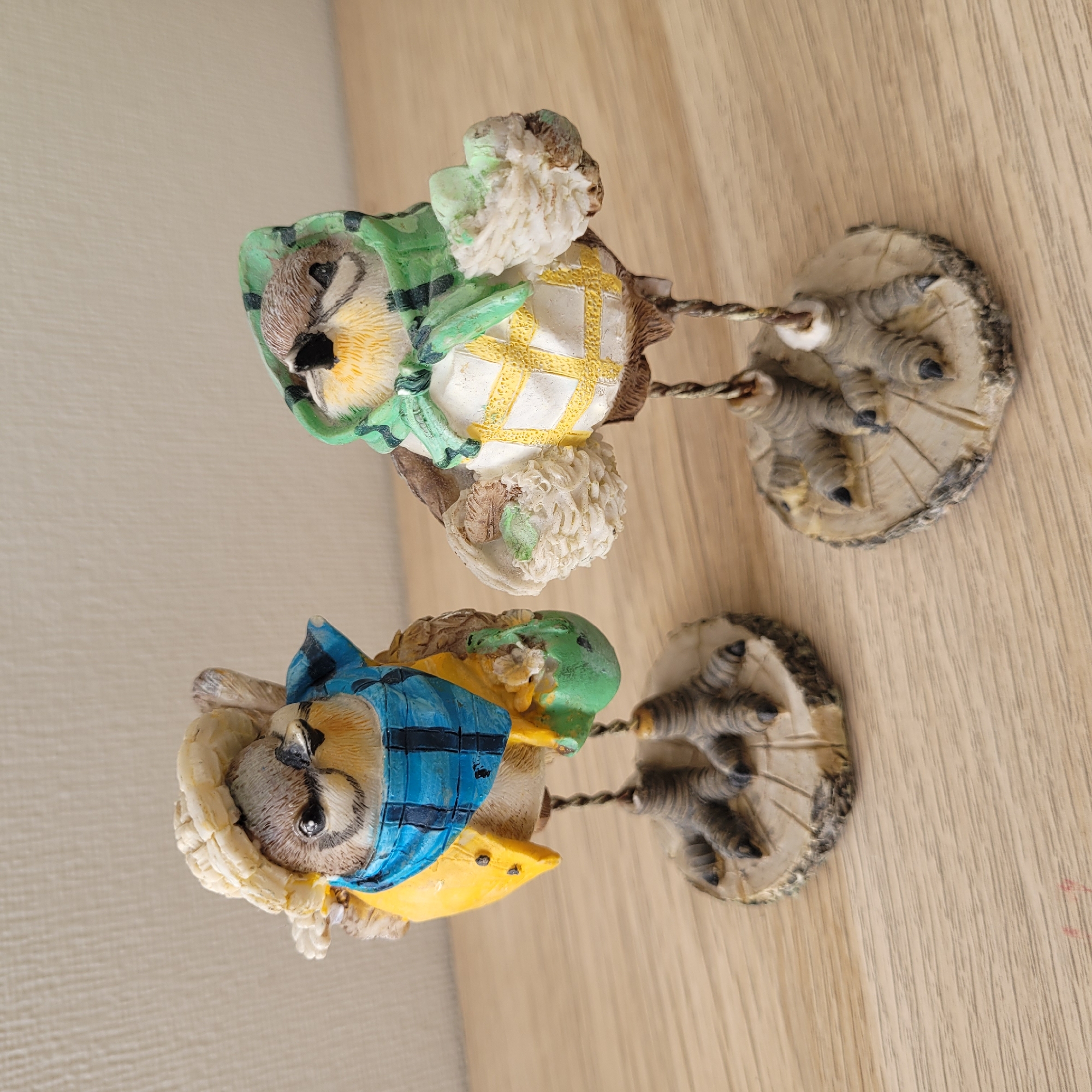 Two ornamental figurines of birds, each standing on a small, textured circular base resembling a tree stump, are positioned on a light-colored wooden surface. The bird on the left wears a straw-like hat, a blue checkered scarf, and a yellow jacket with black buttons. It is holding a small green object. The bird on the right is adorned with a green patterned headscarf and a white garment with yellow criss-cross detailing. It is holding a cluster of white, textured objects. Both birds are supported by thin, twisted metal legs. The background is a plain, textured wall in a light, neutral tone. The image is a close-up, eye-level shot. There is no indication that this scene is located in Jūrmala, Latvia.
