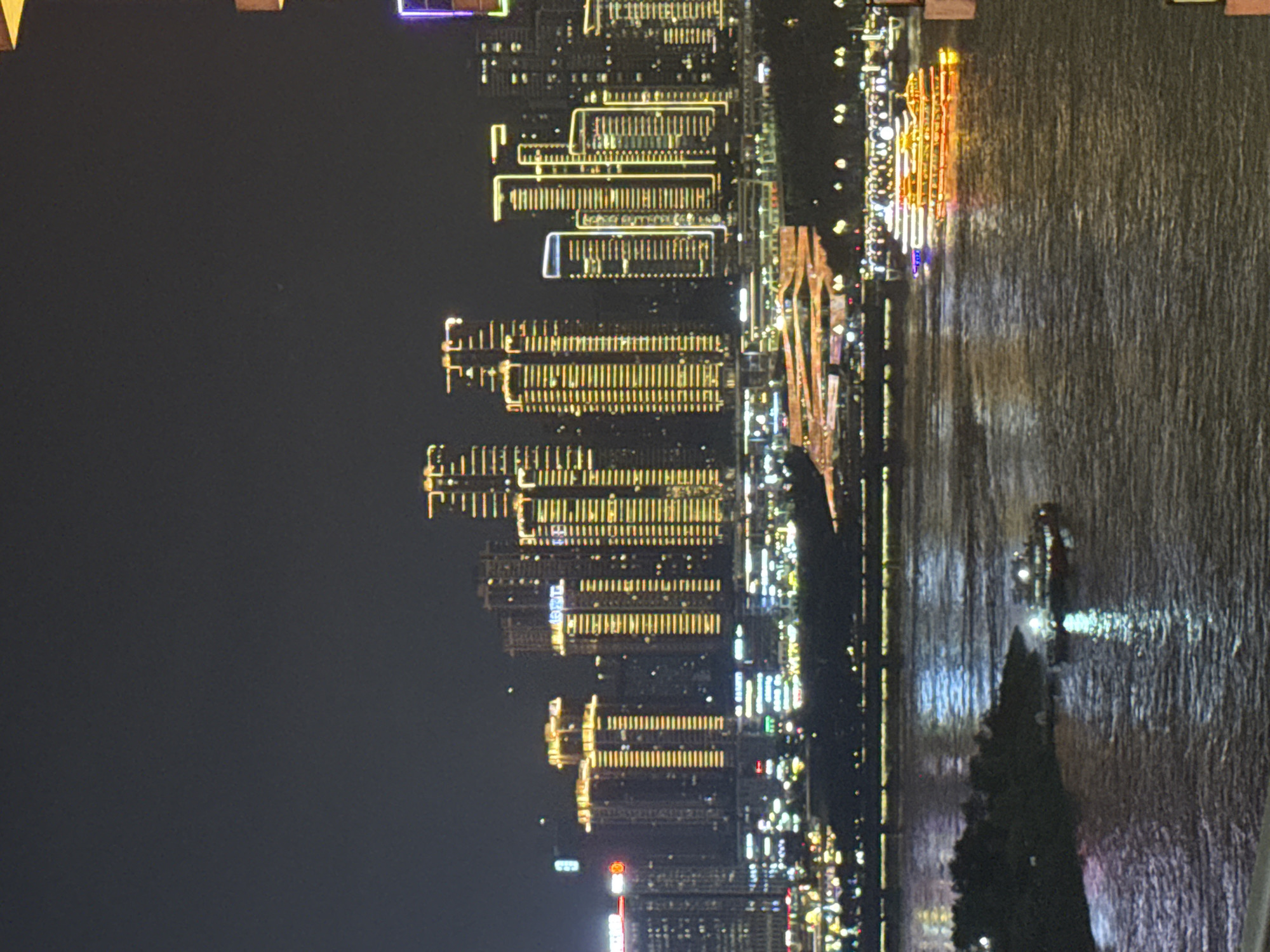 A nighttime, elevated perspective captures a wide river flowing through a city. On the far bank, a dense cluster of modern skyscrapers illuminates the dark sky with a multitude of yellow and white lights, forming an impressive cityscape. Some buildings feature distinct patterns of light outlining their structures. In the foreground, the river's surface reflects the city lights, creating shimmering patterns. Two boats are visible on the water; one in the middle ground is a larger vessel with illuminated decks and an orange glow, while a smaller boat is closer to the left bank, also lit up. The overall scene depicts a vibrant urban environment at night, likely in Yuzhong District, China, known for its riverside development and illuminated skyline.
