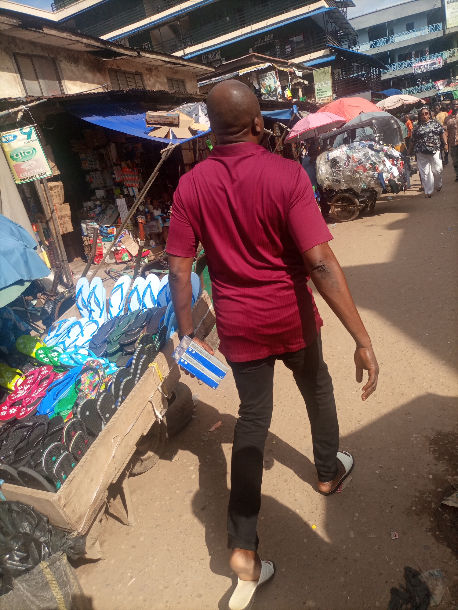 The image depicts a bustling street market scene in Onitsha, Nigeria. In the foreground, a bald Black man wearing a maroon short-sleeved shirt and black trousers walks away from the camera, holding a blue and white rectangular package in his right hand. His left hand is slightly raised, and he is wearing white flip-flops. To his left, a wooden cart is laden with numerous pairs of colorful flip-flops, ranging from bright pink and green to blue and black. Behind the cart, various market stalls with blue tarpaulin roofs are visible, filled with goods, including boxes and what appear to be electronics or household items. Further down the street, other individuals are seen walking, including a woman in a patterned top and white trousers, and a person pushing a cart piled high with various items, including what looks like recycled plastic. The environment is an outdoor market with uneven, dusty ground, and buildings with multiple stories lining the street, some displaying signage. The overall impression is one of a vibrant, commercial area.