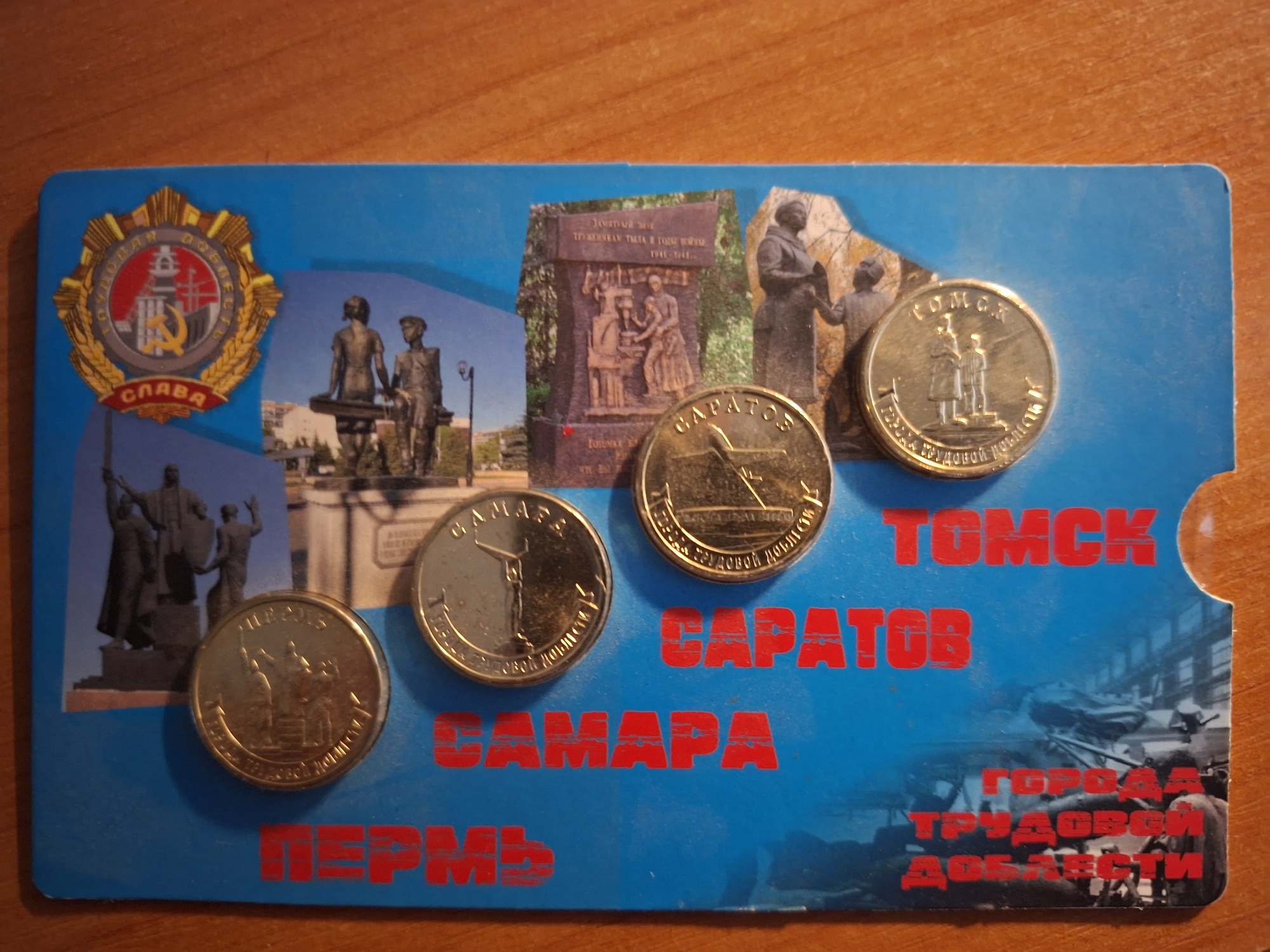 The image shows a blue postcard with several gold-colored coins placed on top of it. The postcard features red text that spells out "ПЕРМЬ," "САМАРА," "САРАТОВ," and "ТОМСК" vertically down the left side. There is also a circular emblem with the word "СЛАВА" in red lettering at the top right of the postcard. Images of monuments and statues are visible on the postcard beneath the coins. The coins themselves appear to be commemorative or collector's items, each bearing different designs and text that is difficult to decipher. The postcard and coins are resting on a wooden surface, likely a table.