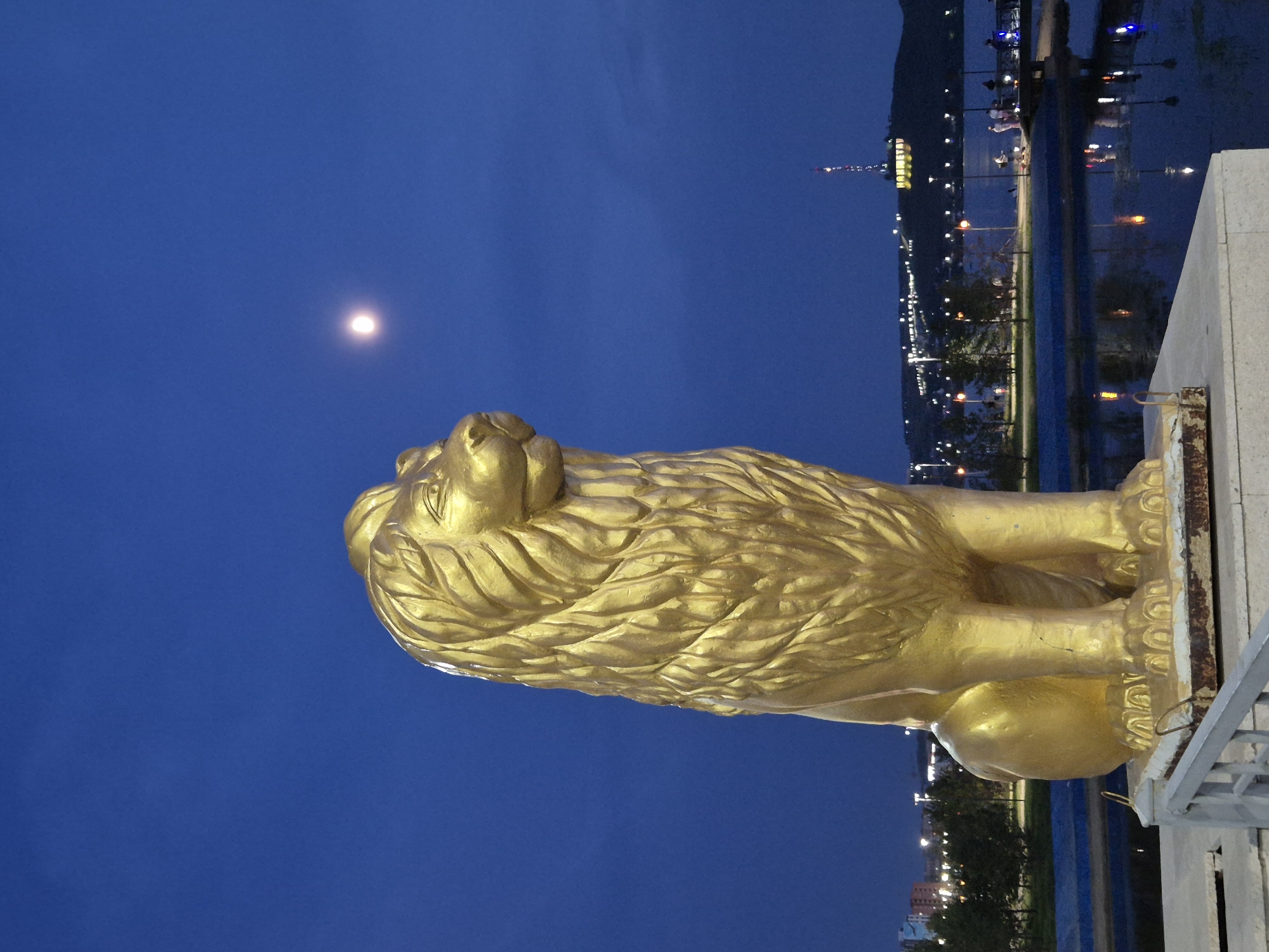 A golden lion statue stands prominently in the foreground, facing to the right. The statue is a monumental sculpture, its surface catching the ambient light. In the background, the night sky is a deep blue, with a faint moon visible in the upper right quadrant. The city of Kokshetau, Kazakhstan, is visible on the horizon across a body of water, which reflects the lights from the city and nearby lamps. Lights from buildings and street lamps dot the hillside, and a tall broadcasting tower with illuminated signage is a distinctive landmark. Several figures are visible on a walkway near the water's edge, some appearing to be fishing. The overall scene suggests an urban park or promenade at dusk or early night.