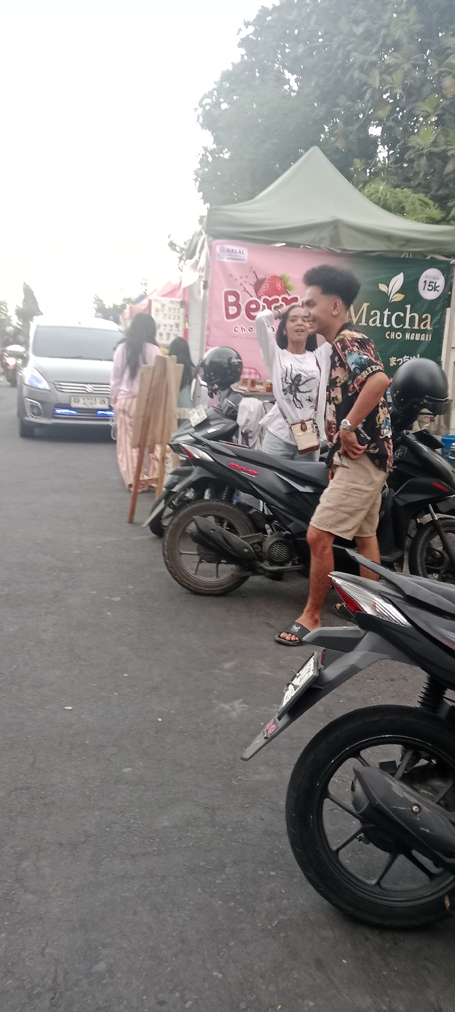 In the city of Ngemplak, Indonesia, a lively street scene unfolds. A young couple, a woman in a white long-sleeved shirt with a spider graphic and a man in a patterned short-sleeved shirt and tan shorts, stand close together, laughing. They are near a food stall with a pink banner advertising "Berry Cho Hawaii" and another green banner with "Matcha Cho Hawaii" and a price of "15k". Several motorcycles are parked in front of the stall. In the background, a silver van is visible, along with other people and tents, suggesting a bustling market or event. The sky is overcast, and the ground is paved asphalt.