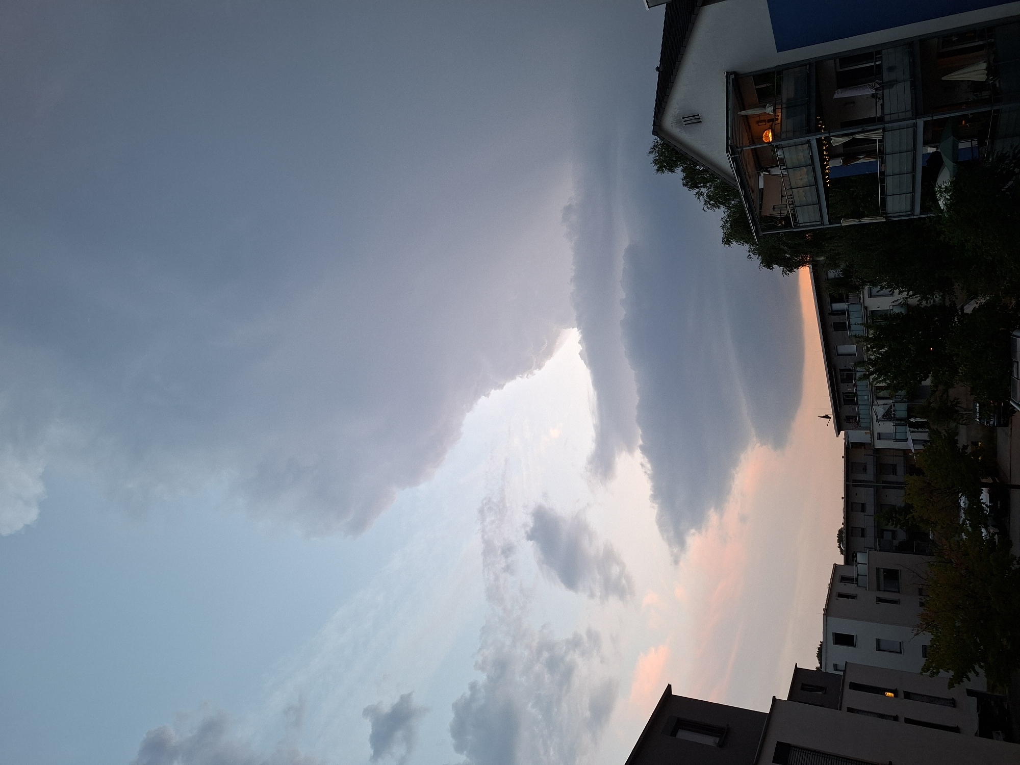 The image shows a cityscape with modern apartment buildings against a dramatic sky filled with large, dark clouds. The sky transitions from a light blue at the top to darker, more ominous clouds towards the horizon, with hints of pink and orange from a sunset or sunrise peeking through. The buildings are contemporary in design, featuring multiple stories, balconies, and windows. Some balconies have furniture and lights visible. Trees are interspersed between the buildings. The overall impression is a cloudy, possibly impending storm in an urban setting. The location is Baden-Baden, Germany. There are no people or animals visible in the image.