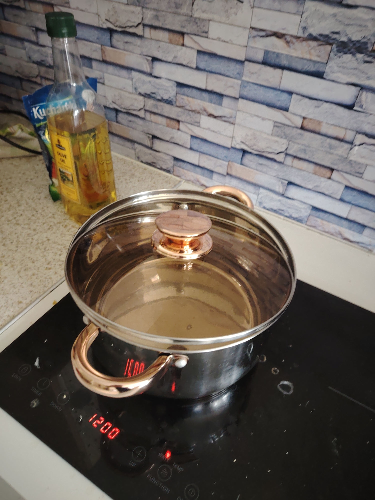 The image displays a kitchen scene with a direct focus on a cooking area. In the foreground, a black induction cooktop is active, showing a red digital display of "1200" on the left burner. Several control buttons are visible, including "LOCK," "TIMER," "DOWN," "UP," "FUNCTION," "WARM," and "TEMP," some illuminated in red. A stainless steel cooking pot with rose-gold colored handles and a matching knob on its clear glass lid is positioned on the active burner; the pot appears to contain a small quantity of clear liquid. Small splatters and debris are visible on the glossy black surface of the cooktop.
To the left of the cooktop, a speckled light-colored countertop holds a clear glass bottle with a green screw cap, containing yellow liquid identified by a partial label as "OLIVE OIL." Behind the bottle, a blue plastic packet, possibly a food item, shows the partially legible word "Kucharia." The background features a tiled wall backsplash, mimicking natural stone or brickwork with rectangular tiles in varying shades of grey, white, beige, and light blue. No individuals are present in the scene. The overall environment suggests a domestic kitchen setting with an ongoing or imminent cooking activity.