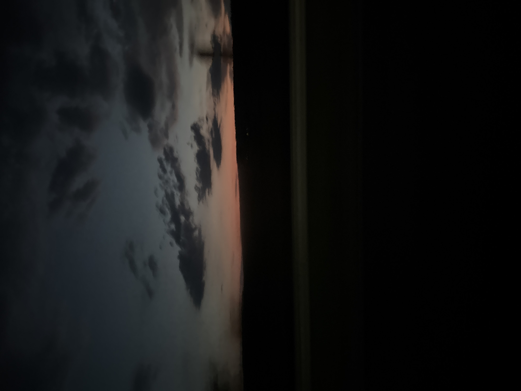 This media file is a photograph of a dramatic sky, likely taken during twilight or sunset. The sky is filled with dark, ominous clouds that are tinged with a warm, peachy-orange hue, suggesting the sun is either rising or setting. These clouds are textured and dynamic, with areas of deep shadow and lighter, illuminated patches.
To the right of the sky, there is a sharp vertical line separating the illuminated clouds from a dark, indistinct area. This dark area appears to be a solid, black void, which could represent a building or a very dark landscape against the brightly lit sky. The transition between the sky and this dark area is abrupt, creating a sense of depth.
The composition is oriented vertically, which emphasizes the vastness and dramatic nature of the sky. The overall mood is somewhat somber and awe-inspiring, characteristic of a powerful sky event. There are no visible people, objects, or text in the image. The scene is likely an outdoor environment, possibly a landscape or cityscape, where the sky is the dominant subject. The time of day is clearly either dawn or dusk due to the color and quality of the light. The weather appears to be cloudy, with the clouds playing a crucial role in the visual impact of the image.