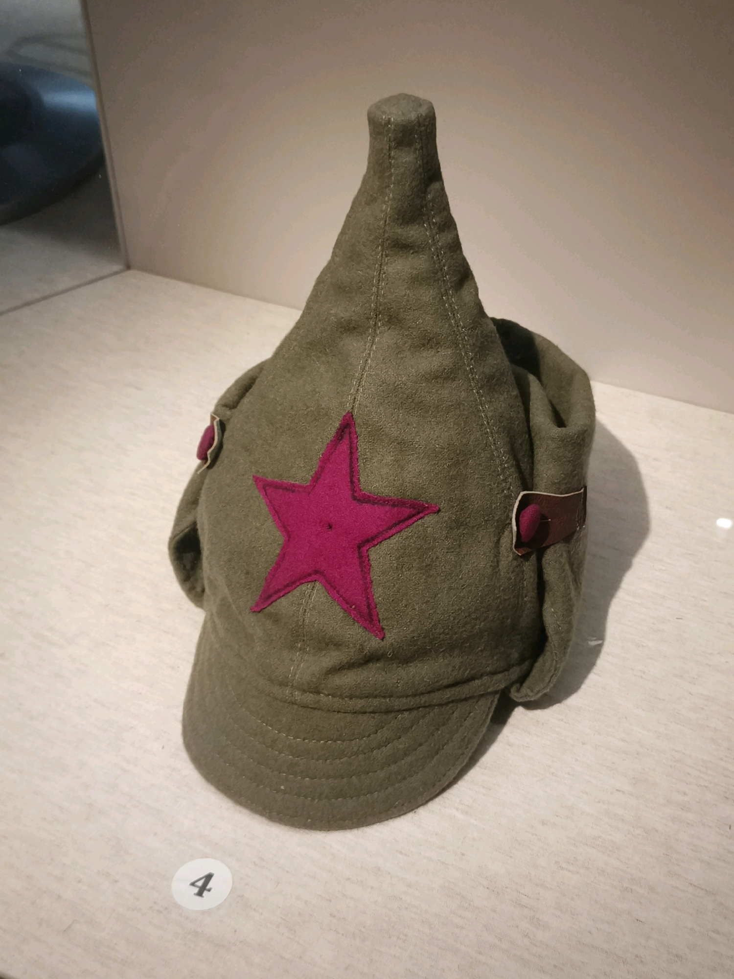 A close-up, eye-level shot shows a khaki-colored military cap with a pointed crown and a brim. A bright pink star is sewn onto the front of the cap. There are leather straps with buttons on either side of the cap. The cap rests on a light-colored, wooden surface. To the left of the cap, a reflection of a dark, rounded object is visible in a mirrored surface. In the bottom left corner of the image, a white circular sticker with the number "4" in black is present. The lighting is even, and the background is a muted, neutral tone, suggesting an indoor display setting, possibly in a museum. The specific location in Cheboksary, Russia, is not discernible from the visual information provided in the image.