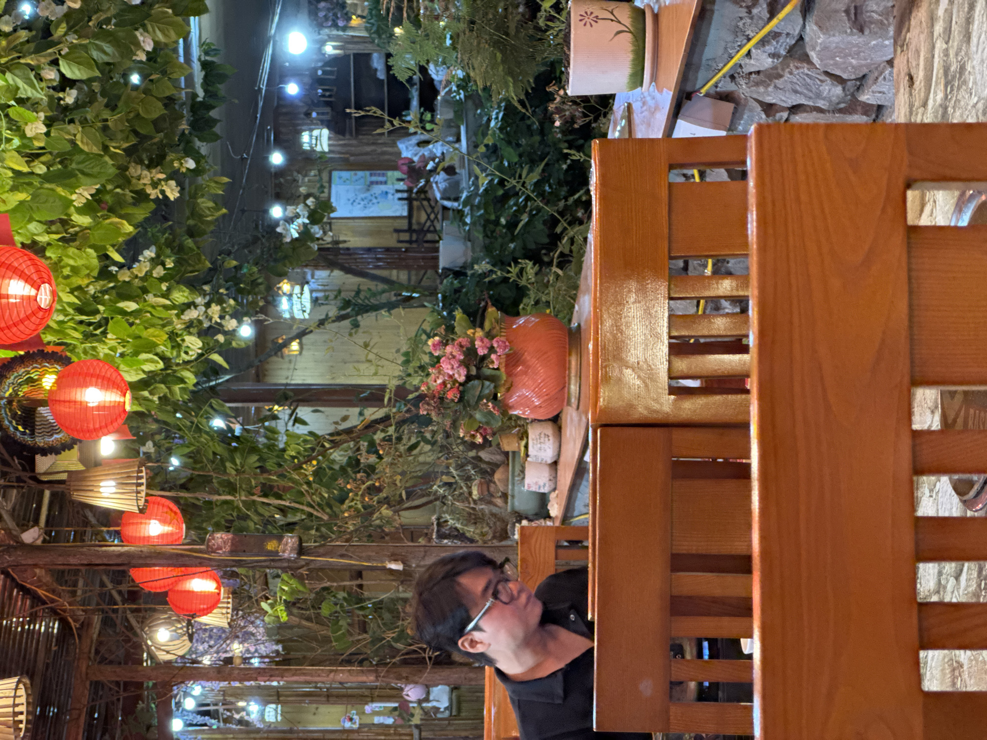 The image depicts a partially obscured view of a person with dark hair and glasses, situated behind a wooden structure, possibly a chair or partition. The individual is wearing a dark shirt and seems to be looking towards the left.
The setting appears to be an outdoor dining area, likely in Mèo Vạc Commune, Vietnam, judging by the foliage, lanterns, and architectural style visible in the background. There are red lanterns hanging amongst lush greenery. Decorative elements, such as potted plants and light fixtures, adorn the area. A path or alley is visible behind the foliage, illuminated by scattered lights. The overall impression is of a lively and inviting outdoor space.