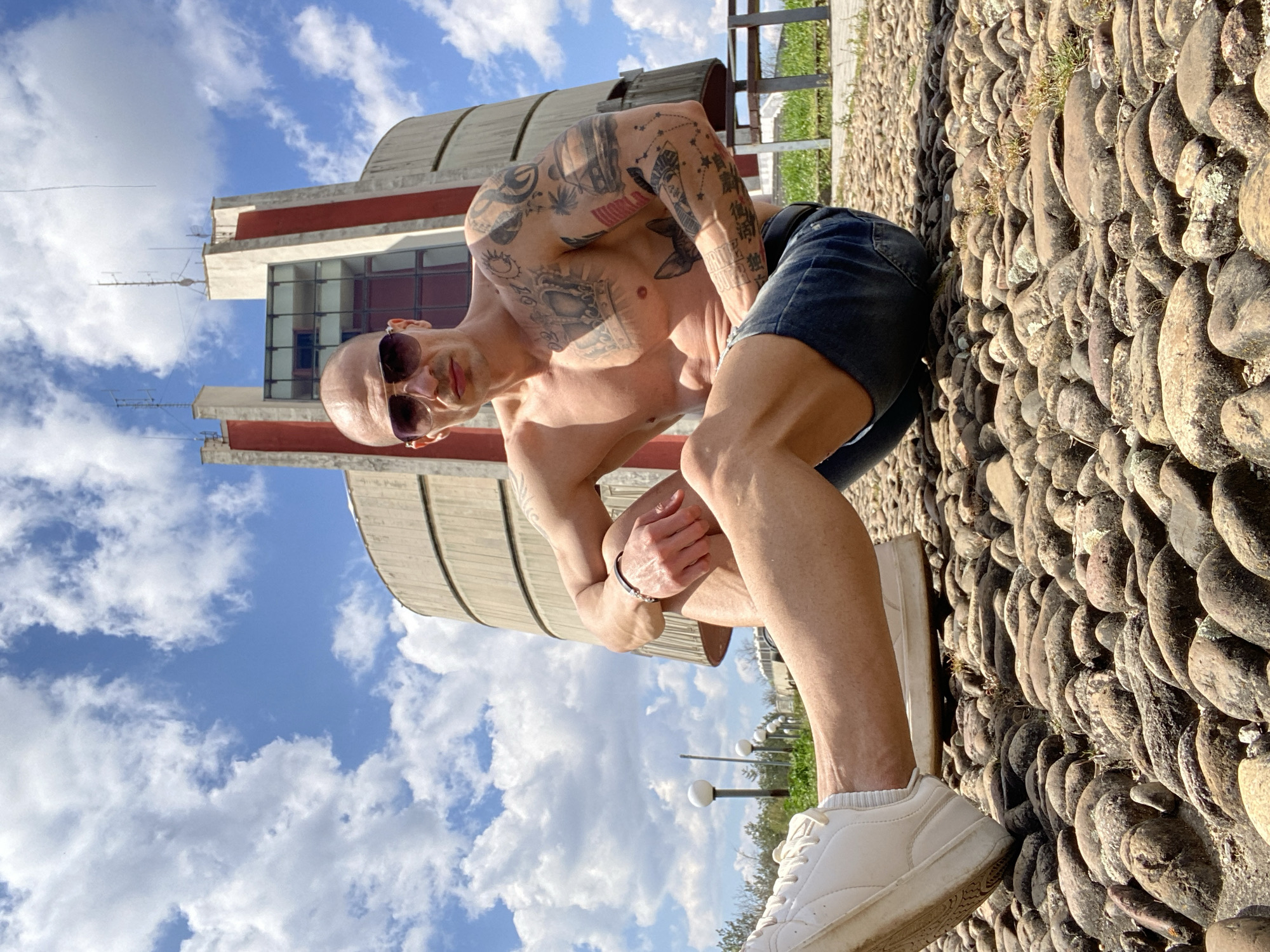 A bald, shirtless man wearing sunglasses and jean shorts stands outdoors. He is tattooed on his arms and chest. He is standing on a walkway made of small, rounded stones, wearing white sneakers. Behind him is a large, cylindrical, concrete building with red accents and rows of windows. The sky is blue with white clouds. Based on the architecture of the building, the location is the Pleven Panorama in Pleven, Bulgaria.