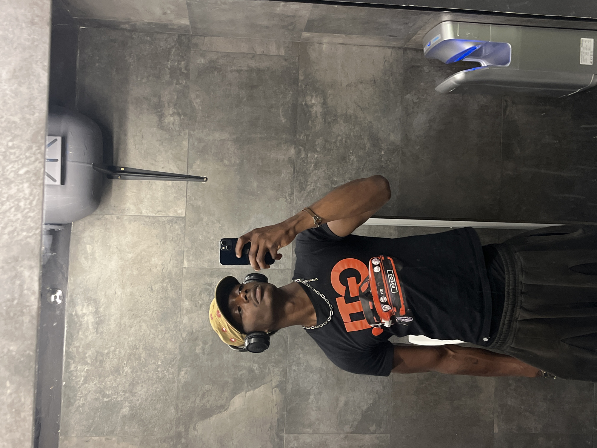 A Black man is taking a selfie in a bathroom. He is wearing a baseball cap, headphones, a black T-shirt with a graphic design, a silver chain, and dark pants. He is holding a smartphone in his right hand. The bathroom has gray tiled walls, a hand dryer, a sink, and a metal towel dispenser. The scene does not definitively indicate Barcelona, Spain. There is no evidence to indicate the location is Barcelona, Spain.