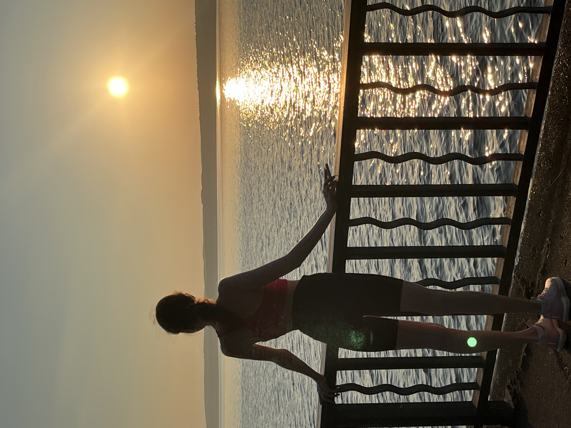A lone individual stands at a railing, looking out at a body of water during sunset. The sun is low on the horizon, casting a warm glow and reflecting brightly off the water's surface. The individual is silhouetted against the sunset, wearing athletic clothing. The railing is black and has a wavy design. The foreground shows a dark, textured surface. The background consists of the water, a distant treeline, and a pale orange sky. The location is the embankment in Chaikovsky, Russia, facing west towards the Kama River.