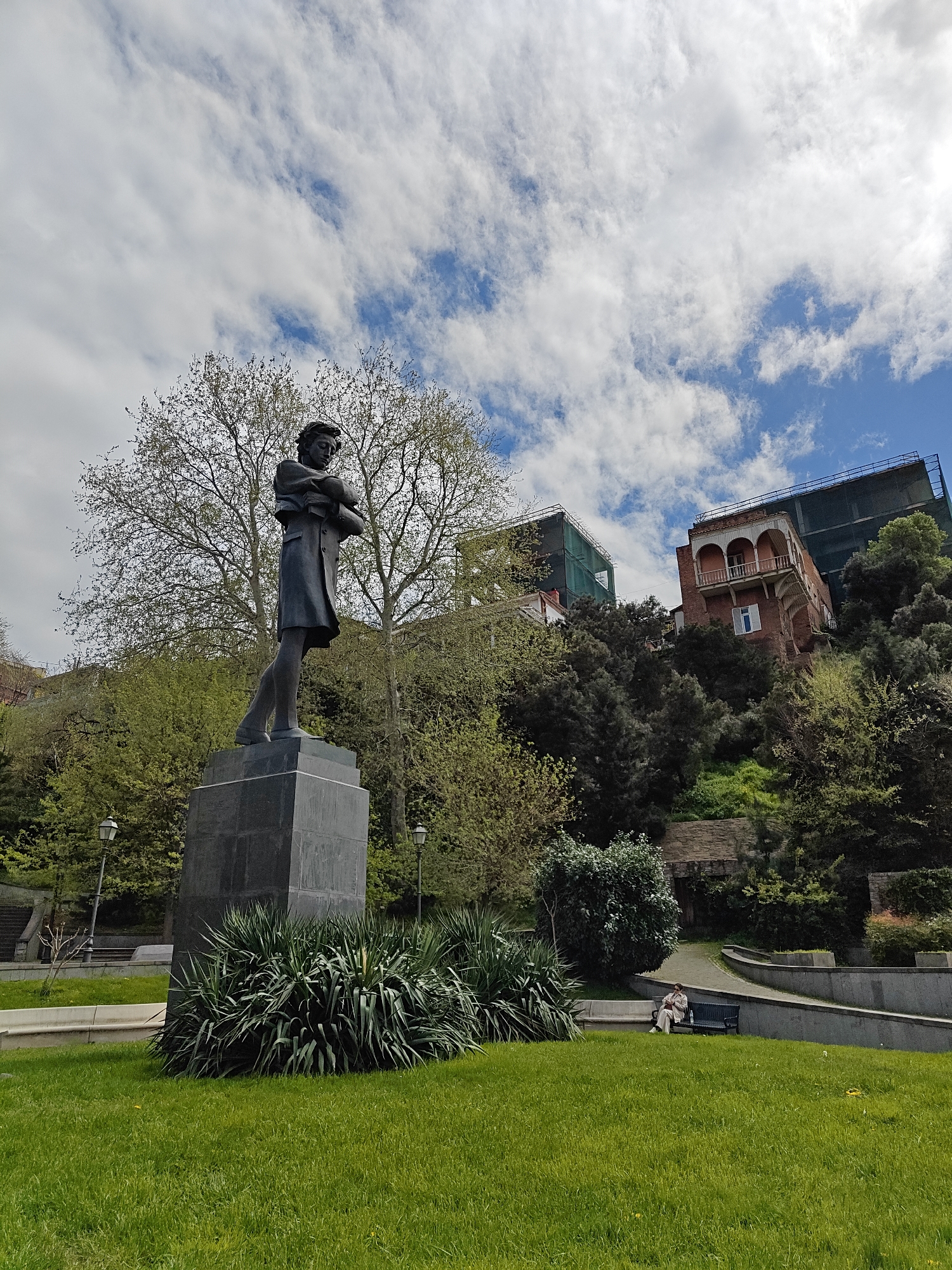 The image displays a daytime scene in a park or public square, likely in Tbilisi, Georgia, under a partly cloudy sky with patches of bright blue.
In the foreground, a tall, dark-colored statue of a man stands on a rectangular dark grey pedestal. The figure is depicted with arms crossed over his chest, a somber expression, and appears to be wearing a long coat. This statue is centrally placed within a well-maintained grassy area. Lush, spiky-leaved bushes are planted at the base of the monument, and vibrant green grass covers the ground in the foreground. Two traditional street lamps are visible near the statue and its base.
The mid-ground features a gently sloping landscape with paved paths and concrete retaining walls, indicative of tiered park design. A variety of trees and shrubs are present; some trees show new spring foliage or bare branches, while others are fully green. To the lower right, a single person is seated on a dark park bench, appearing relaxed and engrossed in something in their hands, possibly a book or phone.
The background reveals a hillside adorned with dense greenery and architectural elements. A prominent red-brick building with arched windows and a decorative balcony stands out, partially covered by scaffolding and green protective netting, suggesting renovation work. Another building with similar scaffolding is visible further to the left on the hillside.
The scene conveys a peaceful atmosphere in an urban green space, blending historical monuments with natural elements and contemporary urban development. No visible text is present in the image.
