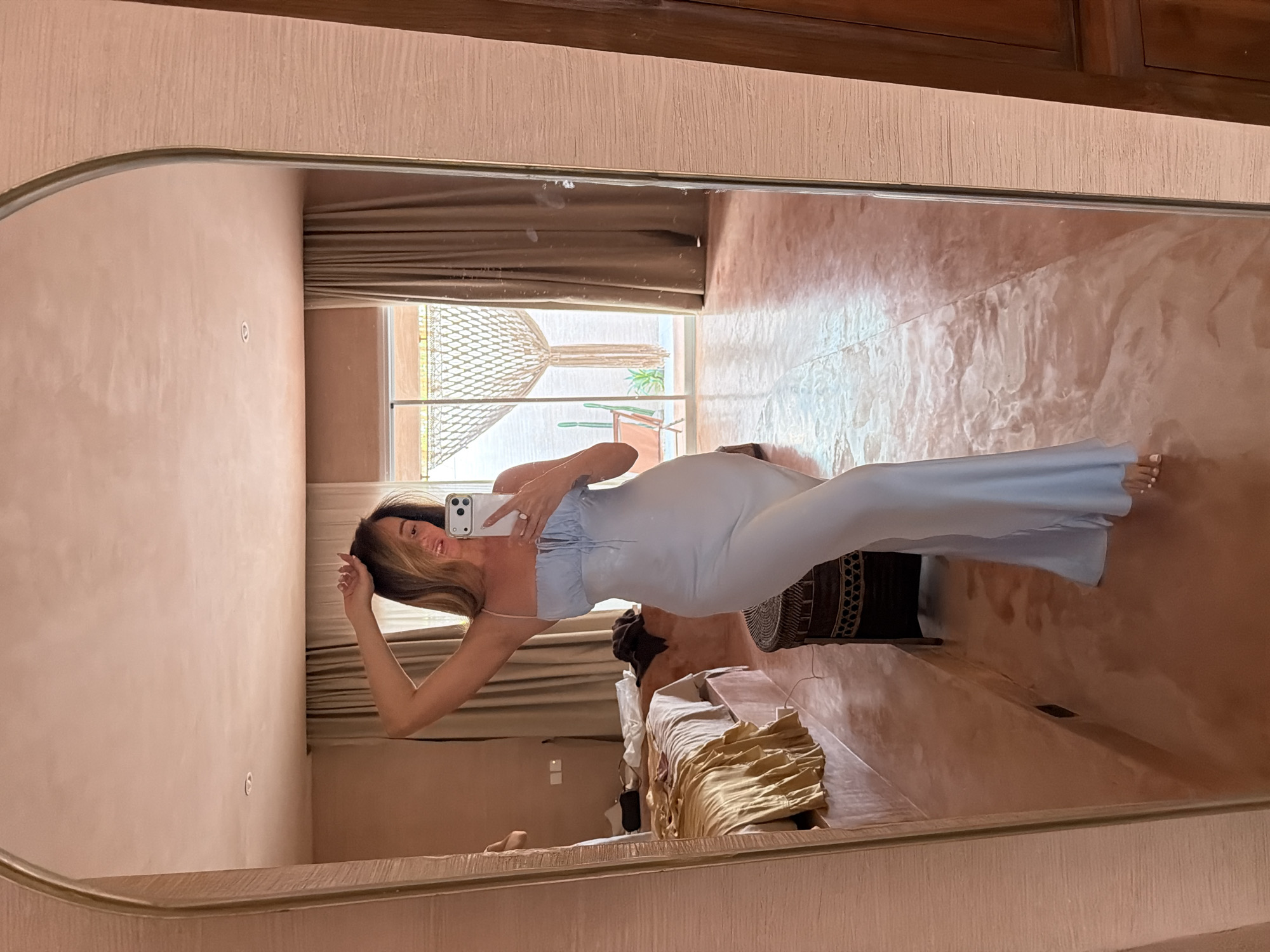 A woman is reflected in a mirror indoors. She is holding a phone in her right hand and posing for a photo. She is wearing a light blue fitted dress. The room has a bed with gold-toned bedding and a brown curtain-like shade. The floor appears to be tiled in a warm, reddish-brown color. Outside the window, there is a view with greenery, possibly indicating a tropical location. The setting suggests a hotel or private residence, possibly located in North Kuta, Indonesia.