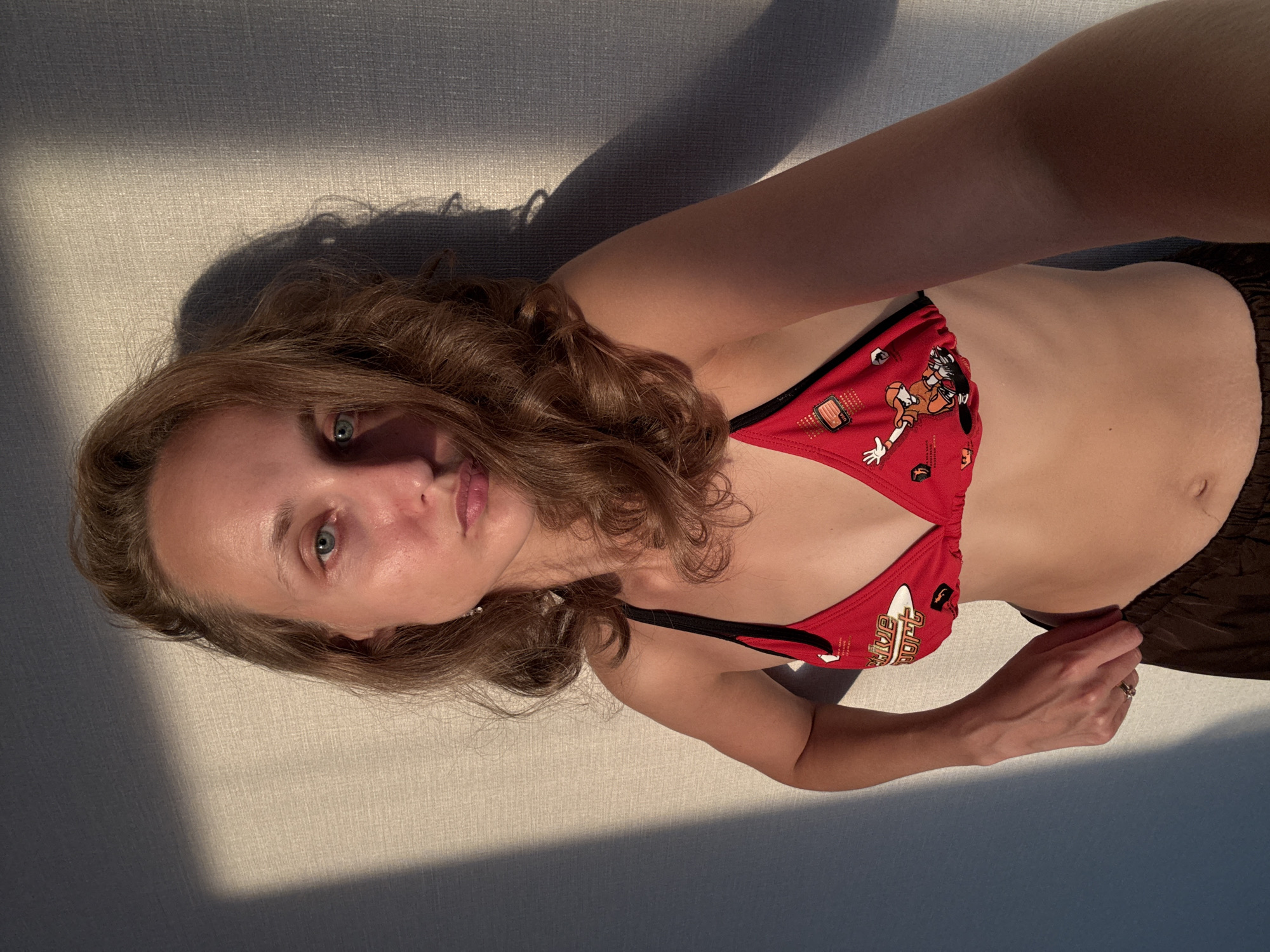 The media file shows a woman lying down, facing the camera. She has light brown hair and blue eyes. She is wearing a red bikini with a cartoon print and brown shorts. The background is a textured wall with sunlight casting shadows, suggesting it is daytime. The lighting is warm and appears to be from a low angle, possibly indicating late afternoon or early morning. The woman's expression is neutral, and she is looking directly at the camera. Her right arm is extended, and she is wearing a ring on her left hand. The overall scene is simple and intimate, with the focus on the woman. There is no discernible text in the image. The context provided indicates the location as Podshivalovo, Russia.