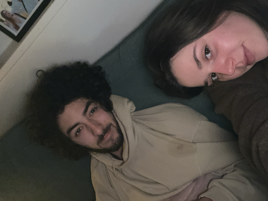 A man and woman are lying down together. The man has dark, curly hair and a beard, wearing a tan hoodie. A stain is visible on the hoodie. The woman has dark hair and is positioned to the right of the man. A framed picture hangs on the wall in the background. They are lying on a blue-green surface, likely a sofa or bed.