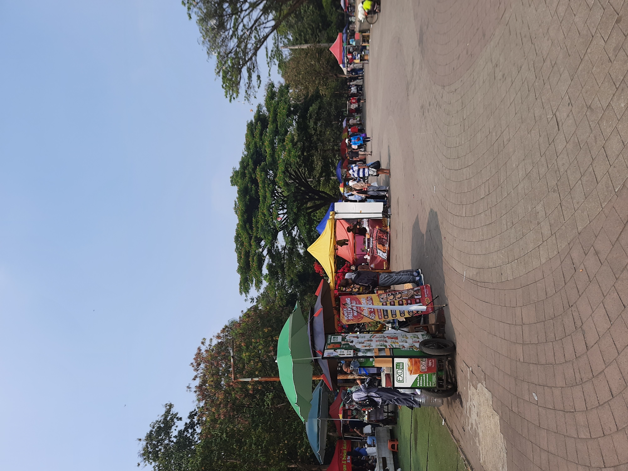 This is a wide shot of an outdoor market on a sunny day. There are several stalls set up, with various food and drinks being sold. The ground is paved with interlocking bricks, and there are trees in the background. There are a number of people browsing the stalls or walking through the market. The overall impression is one of a busy and lively community gathering.