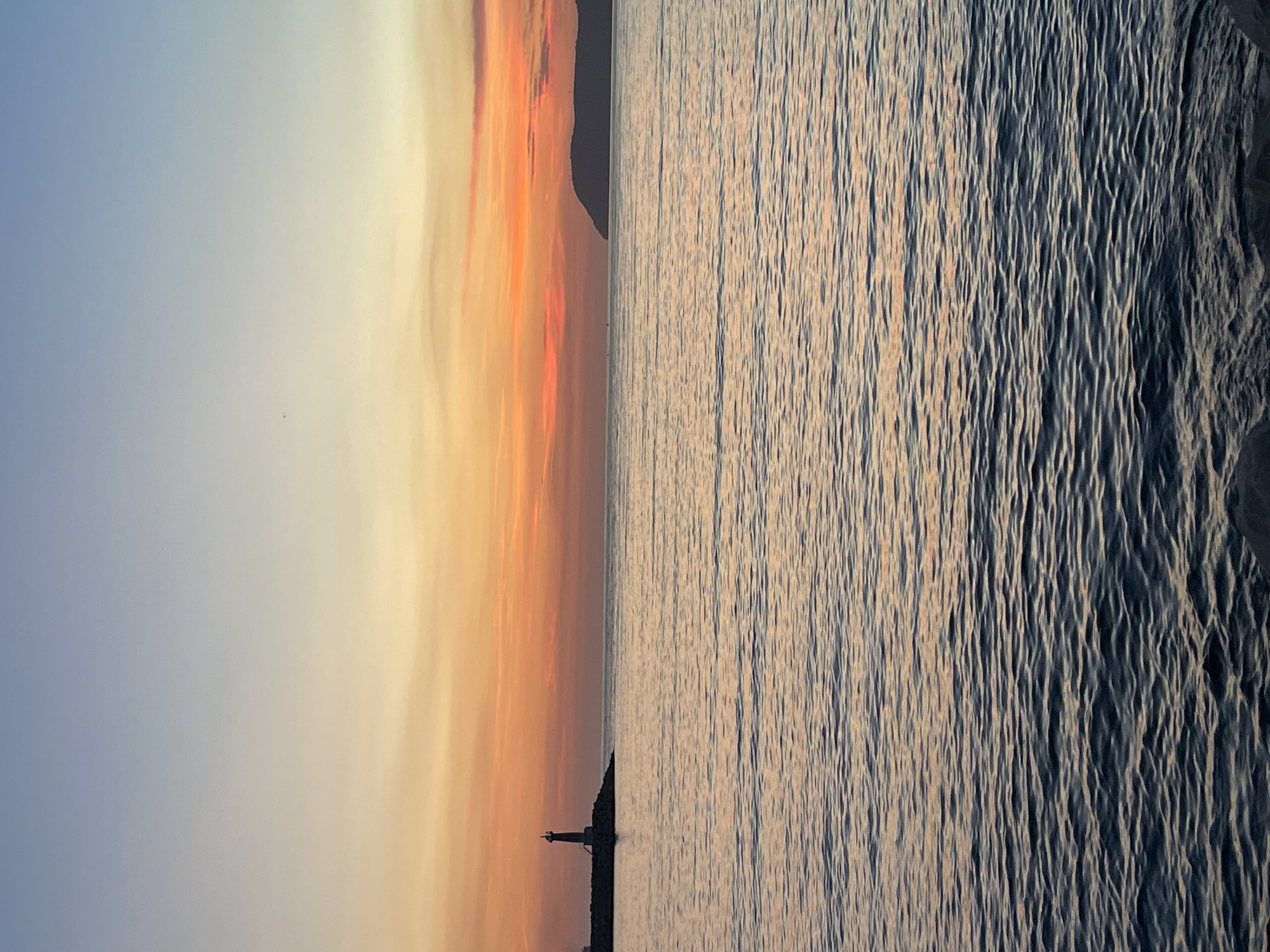 This image captures a serene sunset over the Adriatic Sea, likely near the city of Bar, Montenegro. The sky displays a gradient of colors, transitioning from a soft blue at the top to vibrant shades of orange and red near the horizon, indicative of a sunset.
In the mid-ground, a lighthouse stands tall on a rocky outcrop, silhouetted against the colorful sky. The lighthouse appears to be a solitary structure, marking the edge of the land. To the right, a distant, dark silhouette of a landmass, possibly a peninsula or an island, juts out into the sea.
The sea itself is calm, with gentle ripples reflecting the hues of the sky. The foreground shows the dark, textured surface of the water, with hints of the sunset colors catching the waves. The overall impression is one of tranquility and natural beauty, with man-made elements like the lighthouse adding a touch of human presence to the expansive seascape. There are no people visible in the image, and no actions or interactions are taking place. The environment is natural and coastal.