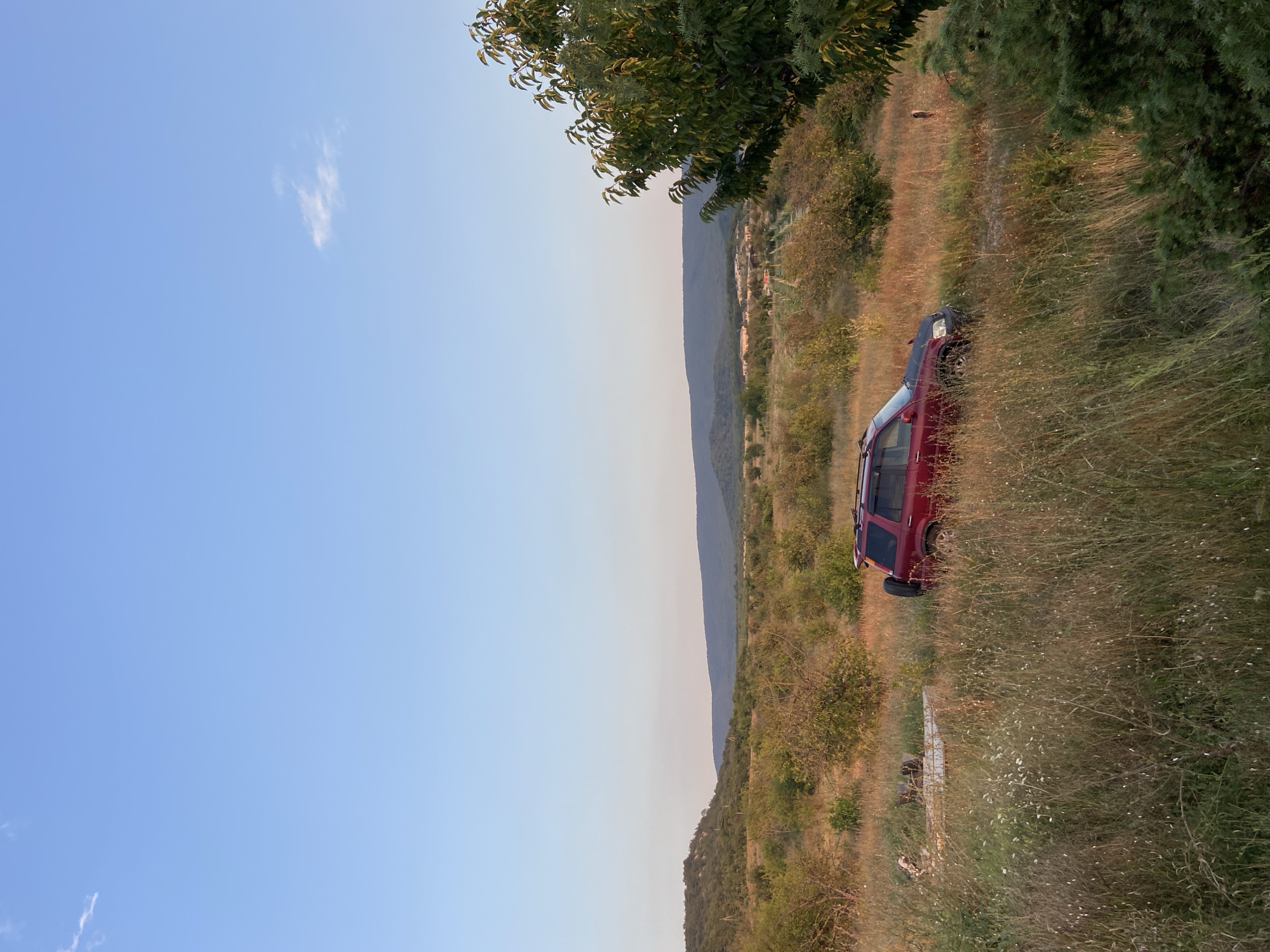 A red SUV is parked in a grassy field with dry, tall grass and scattered shrubs. The vehicle has a roof rack. In the background, rolling hills covered in trees and vegetation are visible, with a clear blue sky above. The scene appears to be in a rural, natural setting, possibly in or around Красномакское сельское поселение, Ukraine.
