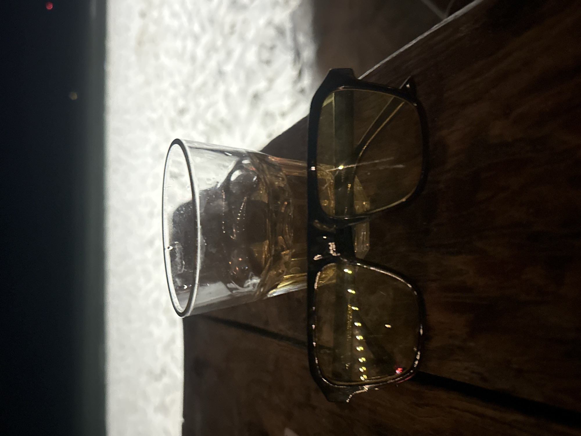 The image depicts a nighttime scene on a dark wooden table, likely outdoors. In the foreground, a pair of dark-framed sunglasses with yellow-tinted lenses rests. Behind the sunglasses, a clear glass tumbler, partially filled with an amber liquid and ice cubes, shows condensation on its exterior. The background features a wide expanse of white, textured surface, identified as sand, stretching towards a dark horizon. Against the dark horizon, faint distant lights are visible: a small yellow point on the left and a red glowing point on the far right. The overall lighting suggests a low-light environment, consistent with nighttime conditions at a beachside location.