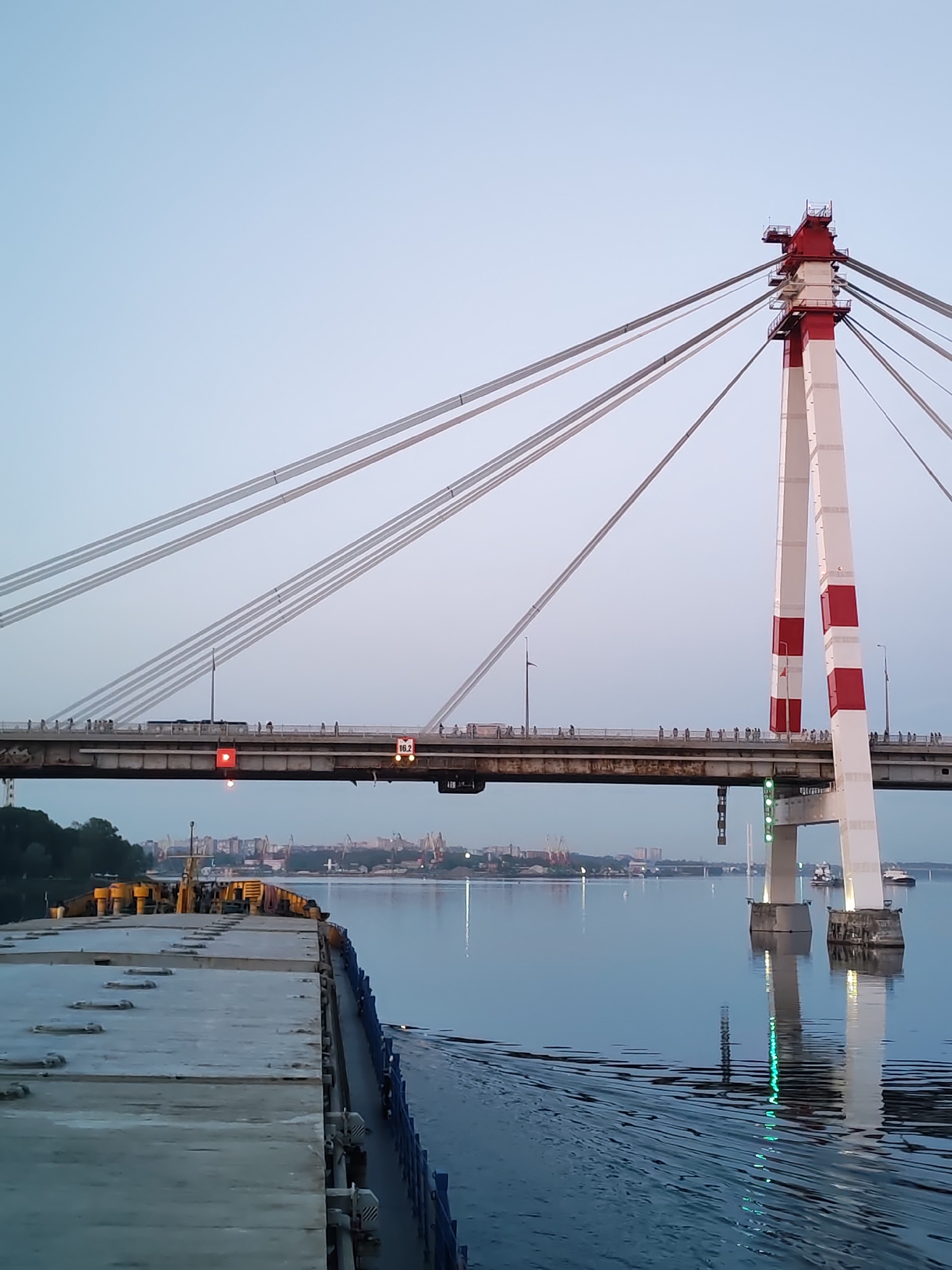 Here is a detailed description of the image:
**Scene Description:**
The image shows a cable-stayed bridge with a red and white support tower, spanning a wide river or waterway. The bridge is populated with a line of people walking across. A barge or large vessel is present in the foreground, situated on the water.
**Objects:**
* **Bridge:** The primary object is a cable-stayed bridge with a distinctive tower. Cables extend from the tower to support the bridge deck.
* **Tower:** The tower has a red and white painted pattern. It is a prominent feature of the bridge.
* **Cables:** Numerous cables support the bridge deck from the tower.
* **Water:** The river or waterway dominates the lower portion of the image. The water surface reflects the surrounding lights.
* **Barge/Vessel:** A large, flat-topped vessel is in the foreground, appearing to be stationary on the water. It is a utilitarian vessel, likely used for transportation.
* **City Skyline:** In the background, a city skyline is visible with various buildings and structures.
* **Lights:** Navigation lights are visible on the bridge and possibly on the barge.
**People:**
* A line of people is walking along the bridge.
**Environment:**
* **Setting:** The scene takes place in an urban environment, as evidenced by the city skyline.
* **Time of Day:** The light suggests either dusk or dawn. The sky is relatively clear.
* **Location:** The location is Unknown, Unknown.
**Interaction:**
* People are walking across the bridge.
* The barge is presumably navigating or moored in the river.