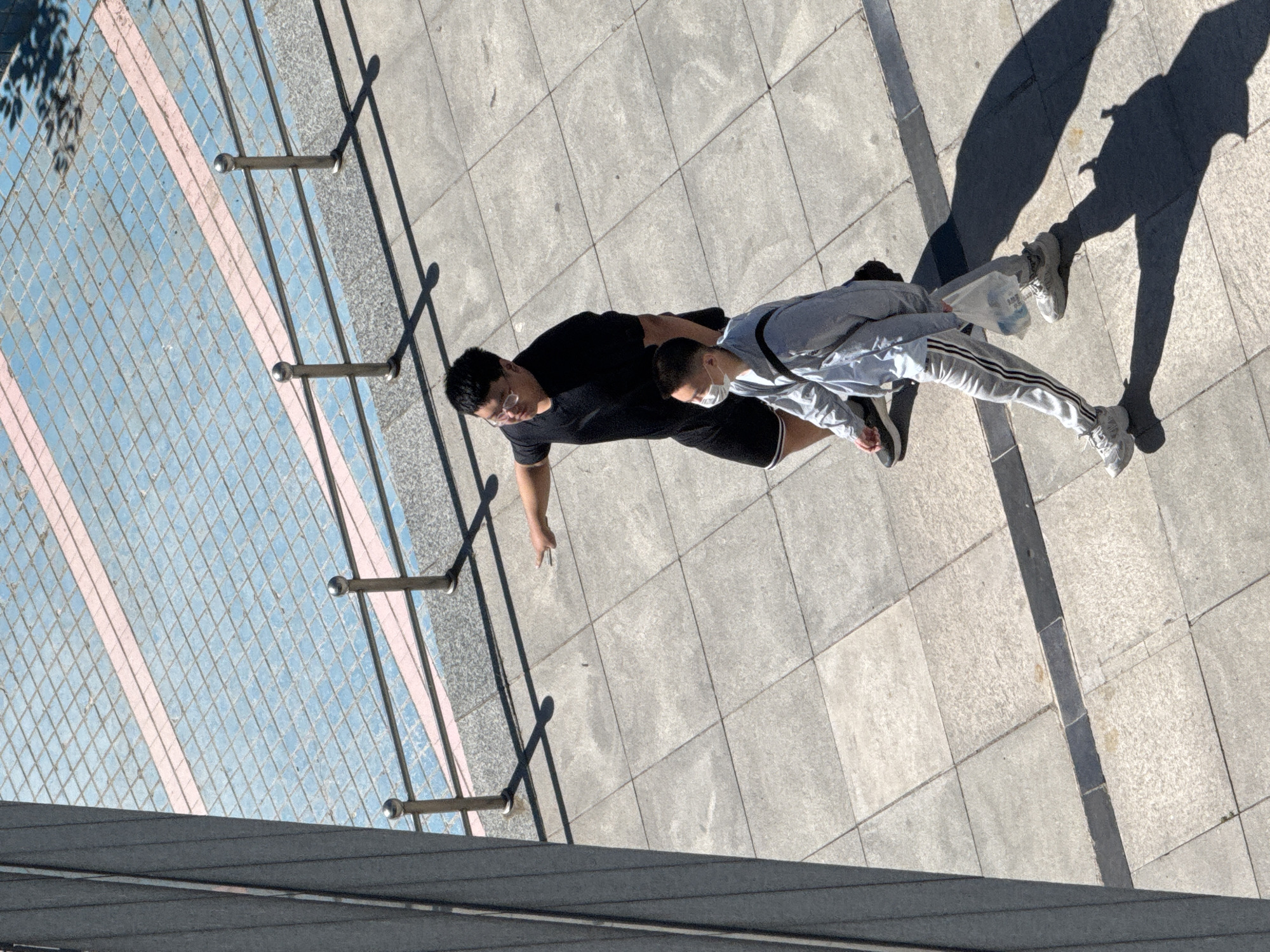 Two individuals are walking across a paved walkway. The person in front is wearing a light grey hooded jacket, light grey sweatpants with black and white stripes on the sides, and white sneakers. This individual is also wearing a white face mask and carrying a clear plastic bag containing water bottles. The person behind is wearing a black t-shirt, black shorts with white trim, and glasses. This person is pointing to their left. The walkway is tiled with large, square grey tiles. Behind the walkway, there is a section with smaller blue tiles and pink stripes. Metal railings with spherical tops are positioned between the tiled sections. A dark, modern building facade is visible on the left side of the image. The scene is illuminated by strong sunlight, casting long shadows. The image was captured from a high vantage point, looking down. This scene is located in Guta, China.