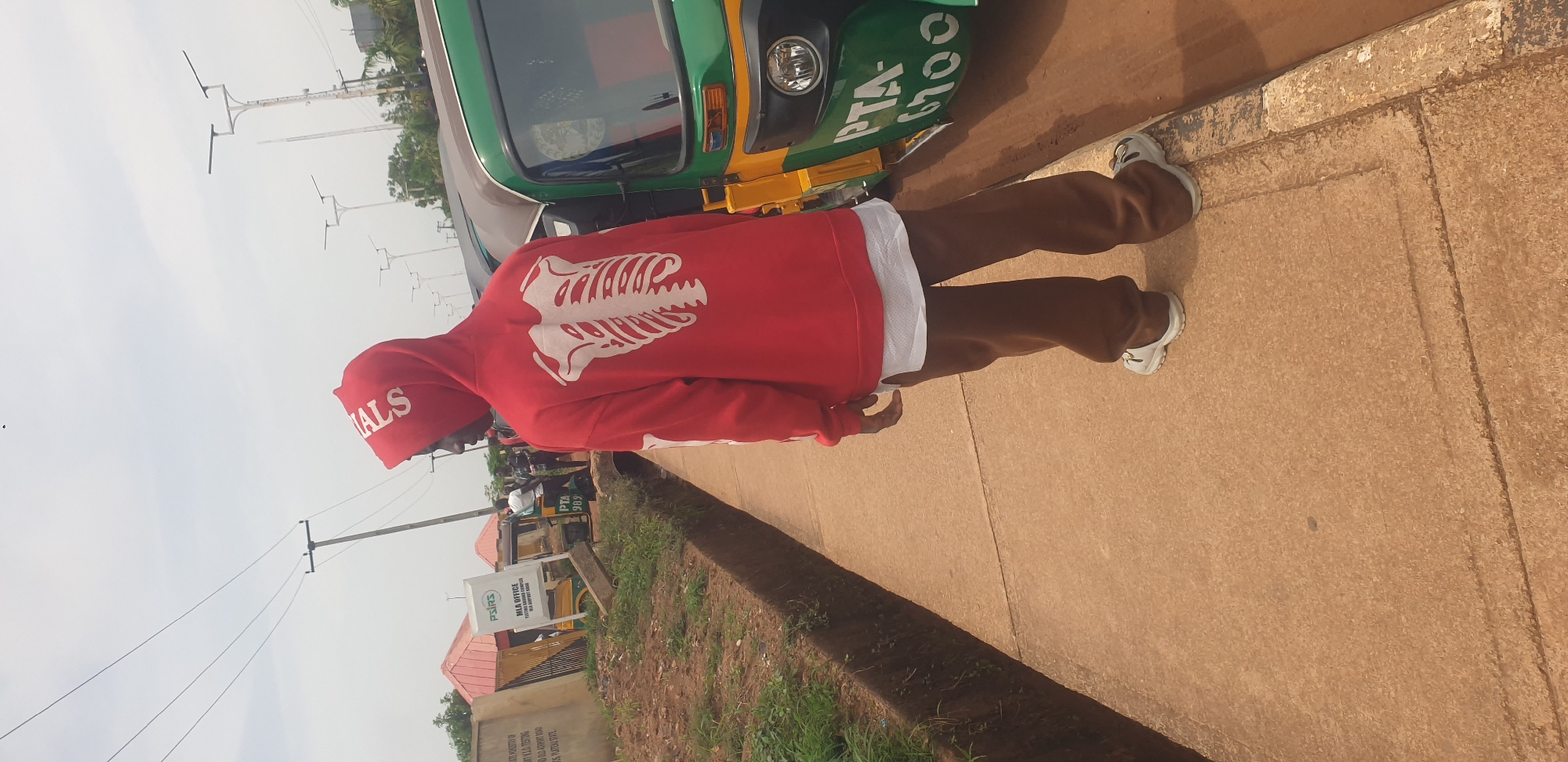A young Black person wearing a red hoodie with a white skeleton design on the back, brown pants, and white sneakers walks along a concrete sidewalk next to a drainage ditch. In the background, a green and yellow tricycle is parked on the side of a dirt road. A white sign with "PTA- 989" is visible on the tricycle. To the left of the person, a fence with a red-roofed building behind it is visible. The sky is overcast. The scene is set in Barakin Lerawi, Nigeria.