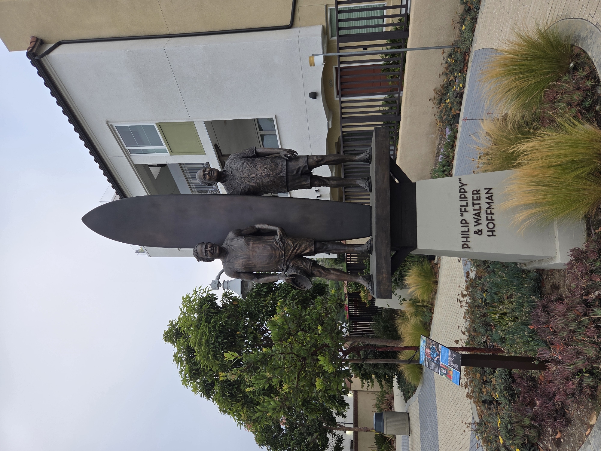 The image shows a bronze statue of two men, Philip "Flippy" and Walter Hoffman, standing next to a surfboard. The statue is located outdoors in Dana Point, California, USA. The men are depicted in casual attire, with one wearing a tank top and shorts and the other a patterned short-sleeved shirt and shorts. Both are wearing sandals. The statue is mounted on a pedestal with the names of the individuals inscribed on it. In the background, there is a modern building with a light-colored facade, balconies, and a tiled roof. A black metal fence runs along the front of the building. The foreground features landscaping with various plants, including ornamental grasses and succulents, and a paved walkway with a decorative pattern. A trash receptacle is visible on the left side of the image, and a sign with information about the individuals is placed near the statue. The sky is overcast.