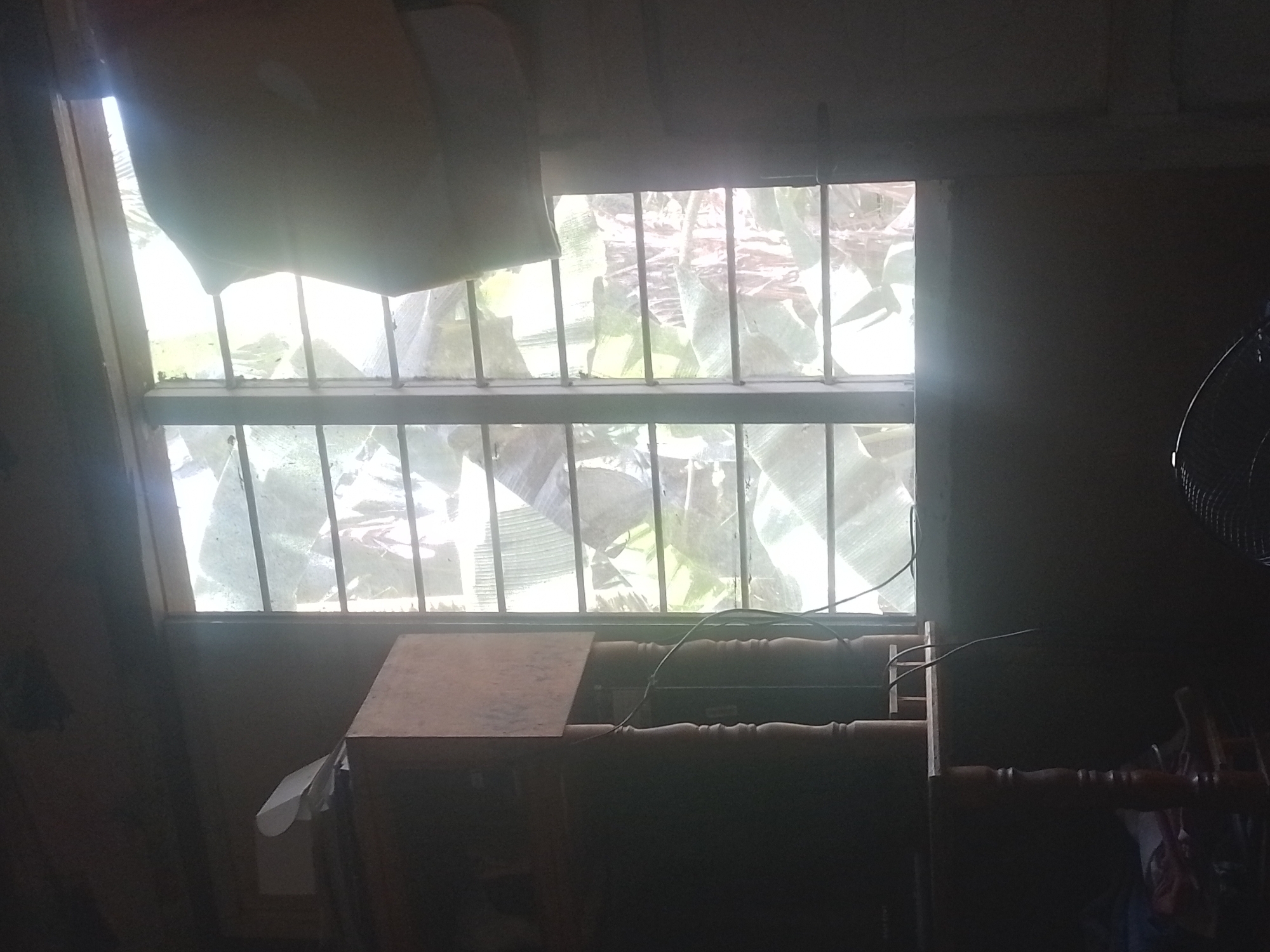 This is a dimly lit indoor scene, likely a residential room in Davao City, Philippines. A window with horizontal bars and a slightly dirty pane dominates the right half of the frame, revealing lush green foliage outside, suggesting a tropical environment. Sunlight streams through the window, creating a bright, almost blown-out effect in some areas and casting faint shadows.
To the left of the window, a wooden cabinet or shelf unit is visible. On top of it, there are several papers, one of which is curved upwards. Part of a television screen is also visible on the cabinet. Below the cabinet, a portion of a wooden table with turned legs is present, and some fabric items are strewn on the floor beneath it. A black electrical wire snakes down from the cabinet area and across the wall. In the bottom right corner, the rounded metal grille of an electric fan is partially visible. The overall impression is of a simple, somewhat cluttered living space.