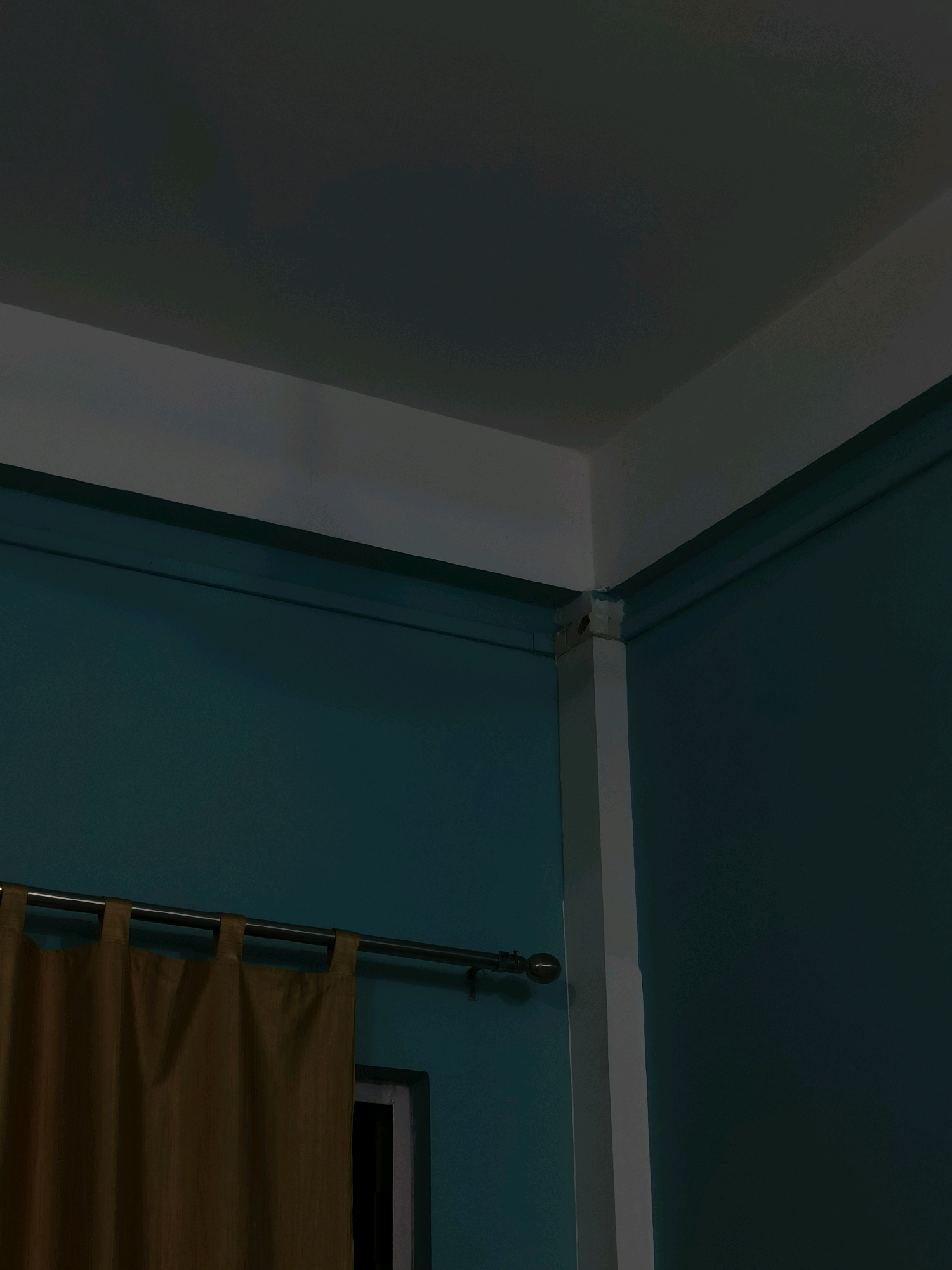 This is a low-angle, close-up, indoor photograph of an interior corner of a room. The walls are painted a dark teal color, and the ceiling is a lighter, off-white. A white corner molding runs vertically along the corner where the two walls meet, extending up to the ceiling. A dark brown curtain hangs from a silver rod on the left side of the frame, obscuring a portion of the wall and a sliver of what appears to be a window frame. The lighting is dim, casting shadows and giving the scene a muted appearance. The location of Doimukh, India, is not discernible from the visual elements present in the image. There are no people or actions depicted.