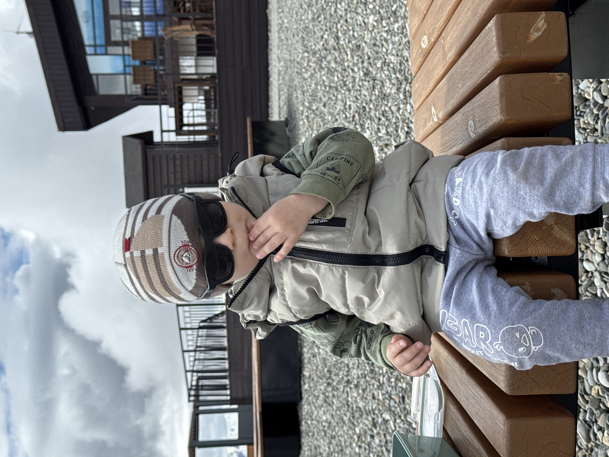 A toddler wearing sunglasses and a knit hat, light grey jacket, olive green shirt, and grey pants with a white koala graphic is reclining on a wooden bench. The bench is made of horizontal wooden planks. Behind the bench is a gravel area, and behind that is a building with glass windows and a dark brown facade. The sky is cloudy. The photo was taken in Estosadok, Russia.