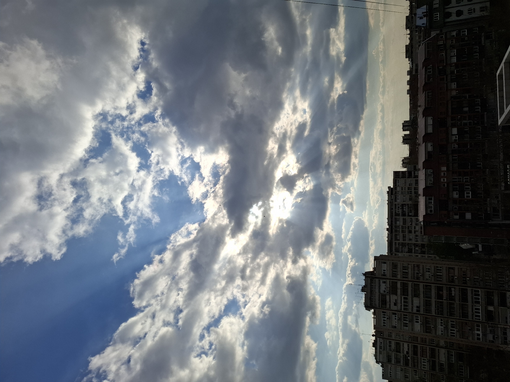 The image shows a partly cloudy sky over Novi Sad, Serbia. The sky transitions from a clear blue to layered clouds with the sun peaking through. The right side of the image includes multiple tall residential buildings. There are no visible people in the image.