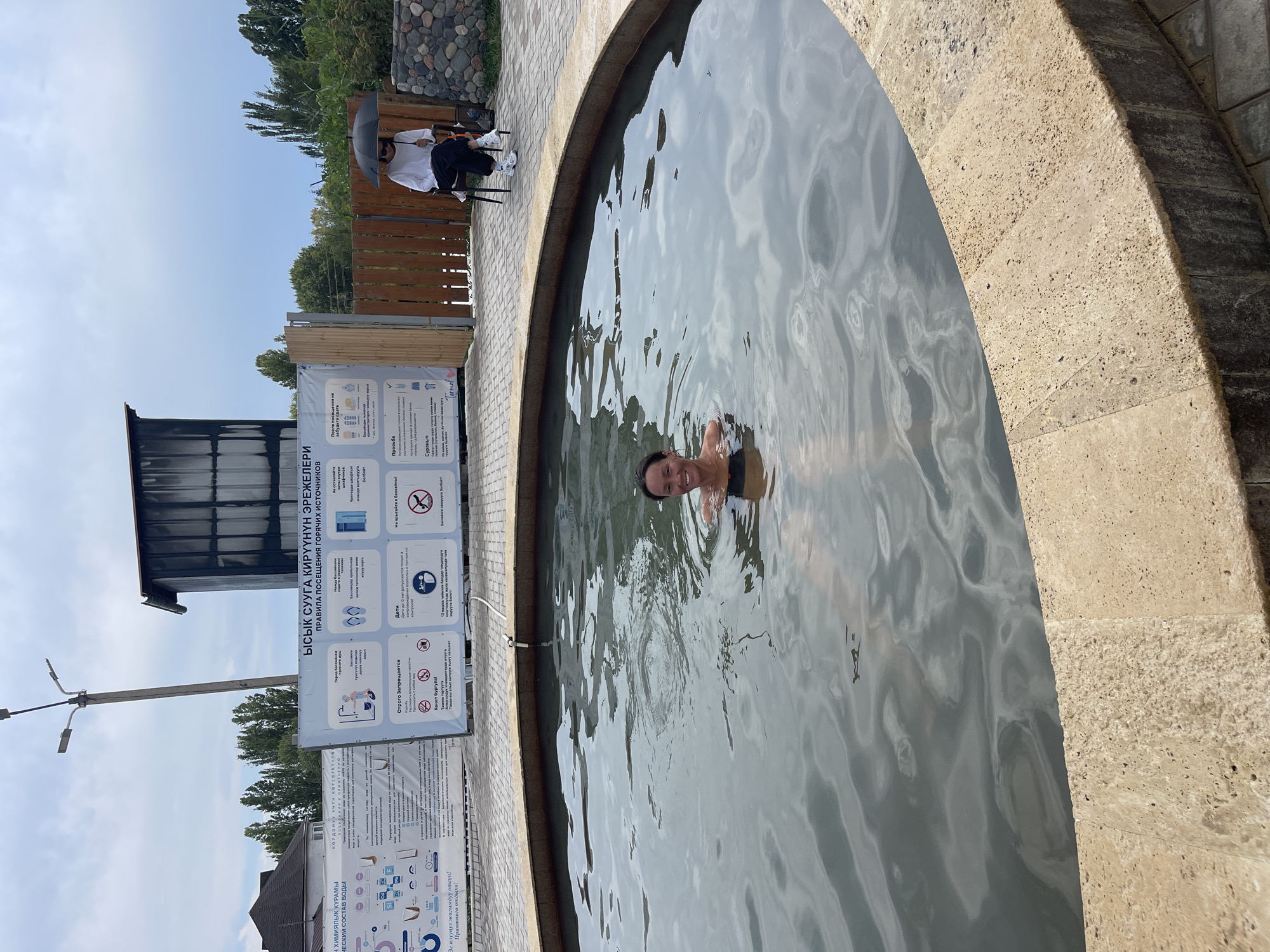 A woman is partially submerged in a circular, shallow pool of dark water, smiling at the camera. The pool has a tan, textured edge and is surrounded by grey pavers. In the background, to the right, a person sits on a white chair holding a black umbrella over their head, facing away from the camera. Behind them, a wooden fence encloses the area, and beyond that, a stone wall and trees are visible. To the left, large informational posters are displayed on white boards, featuring text and graphics in what appears to be Cyrillic and possibly Kazakh. A tall lamppost with multiple lights stands near the posters. The sky above is overcast. The location is identified as Cholpon-Ata, Kyrgyzstan.