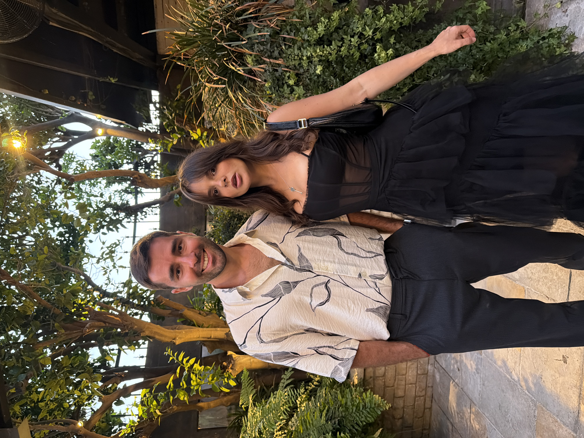 A man and a woman pose in front of vegetation. The man has short brown hair and is wearing a light-colored, patterned, short-sleeved shirt and dark pants. He is smiling. The woman is wearing a black dress with sheer details and a black handbag. Her hair is long and brown. They are standing close together, and the woman's arm is raised. The environment includes trees, greenery, and a brick wall. The location appears to be an outdoor setting in Tel-Aviv, Israel. The lighting suggests it may be late afternoon or early evening.
