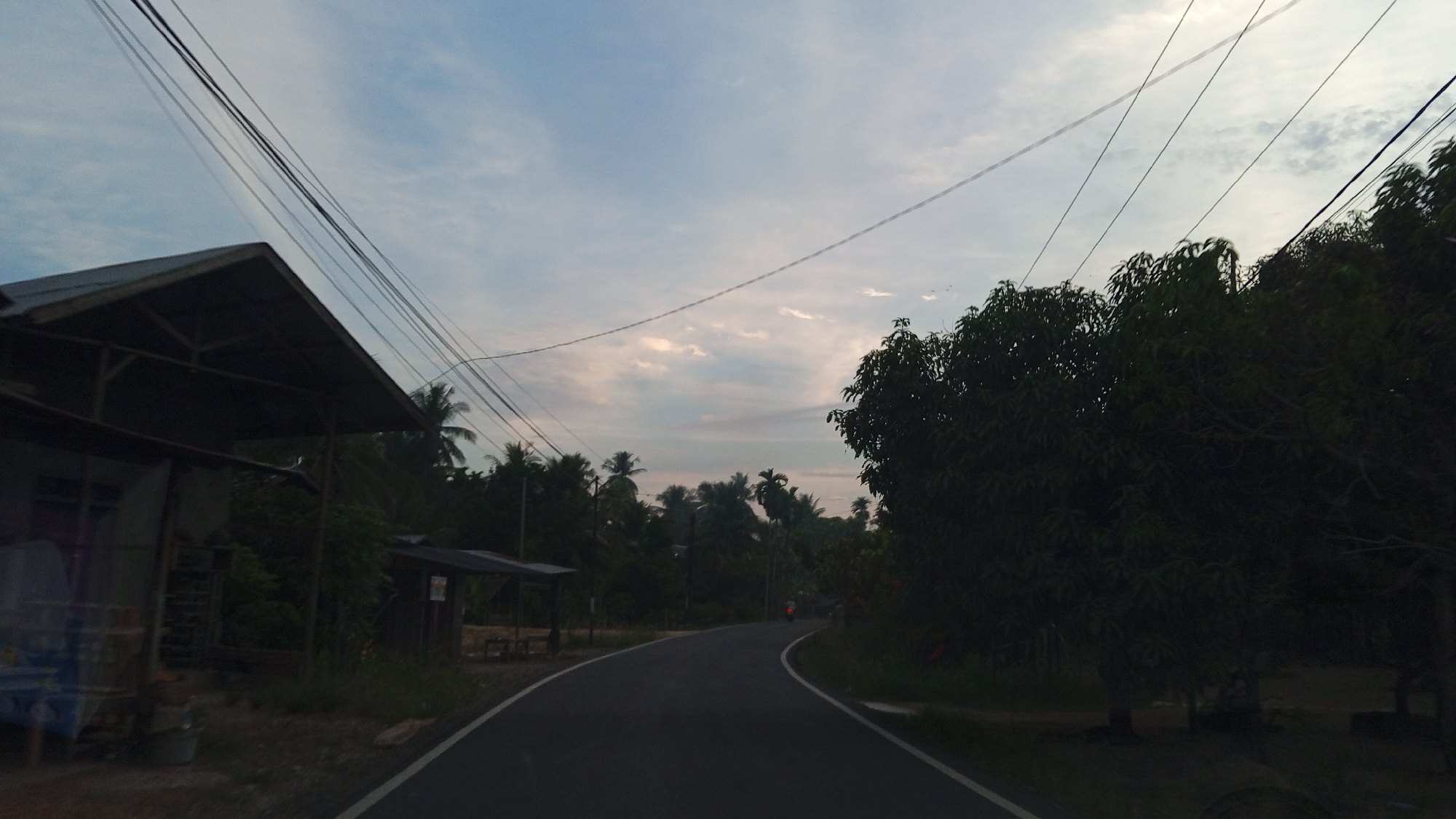 The image shows a rural road in Luengbaro, Indonesia, at dusk. The sky is a pale blue with scattered clouds, and power lines stretch across the frame. On the left side of the road, a small, weathered building with a dark, pitched roof is visible, possibly a roadside stall or dwelling. Further down the road, another small, open-sided shelter with a dark roof stands beside the road. Lush green trees, including palm trees, line both sides of the road, creating a dense foliage. A dark-colored motorcycle with a rider is visible on the road, moving away from the viewer and turning a bend. The road itself is paved and has white lines marking its edges. The overall impression is of a tranquil, natural setting on the outskirts of a village.