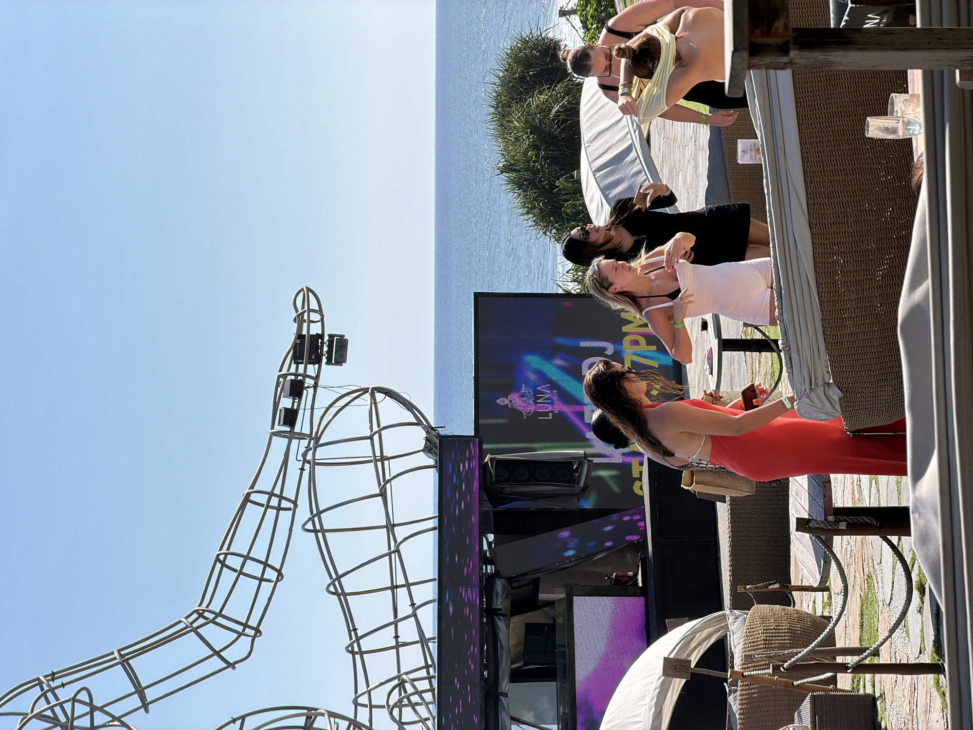 In Kediri, Indonesia, a group of women are gathered outdoors by the sea. In the background, a large metal sculpture structure is visible against a clear blue sky. Below it, a stage is set up with screens displaying colorful lights and a logo for "LUNA BEACH CLUB."
In the foreground, four women are engaged in various activities. One woman in a bright red dress stands near a table, holding a phone and looking down. Next to her, another woman in a white outfit adjusts her clothing. A third woman in a black dress looks away from the camera, while the fourth woman, in the foreground and partially obscured, is seen pulling a yellow garment over her head.
The environment appears to be a beach club or resort, with wicker furniture, lounge chairs, and plants. The ocean is visible in the distance, with gentle waves. The overall scene suggests a relaxed, social gathering in a tropical setting.