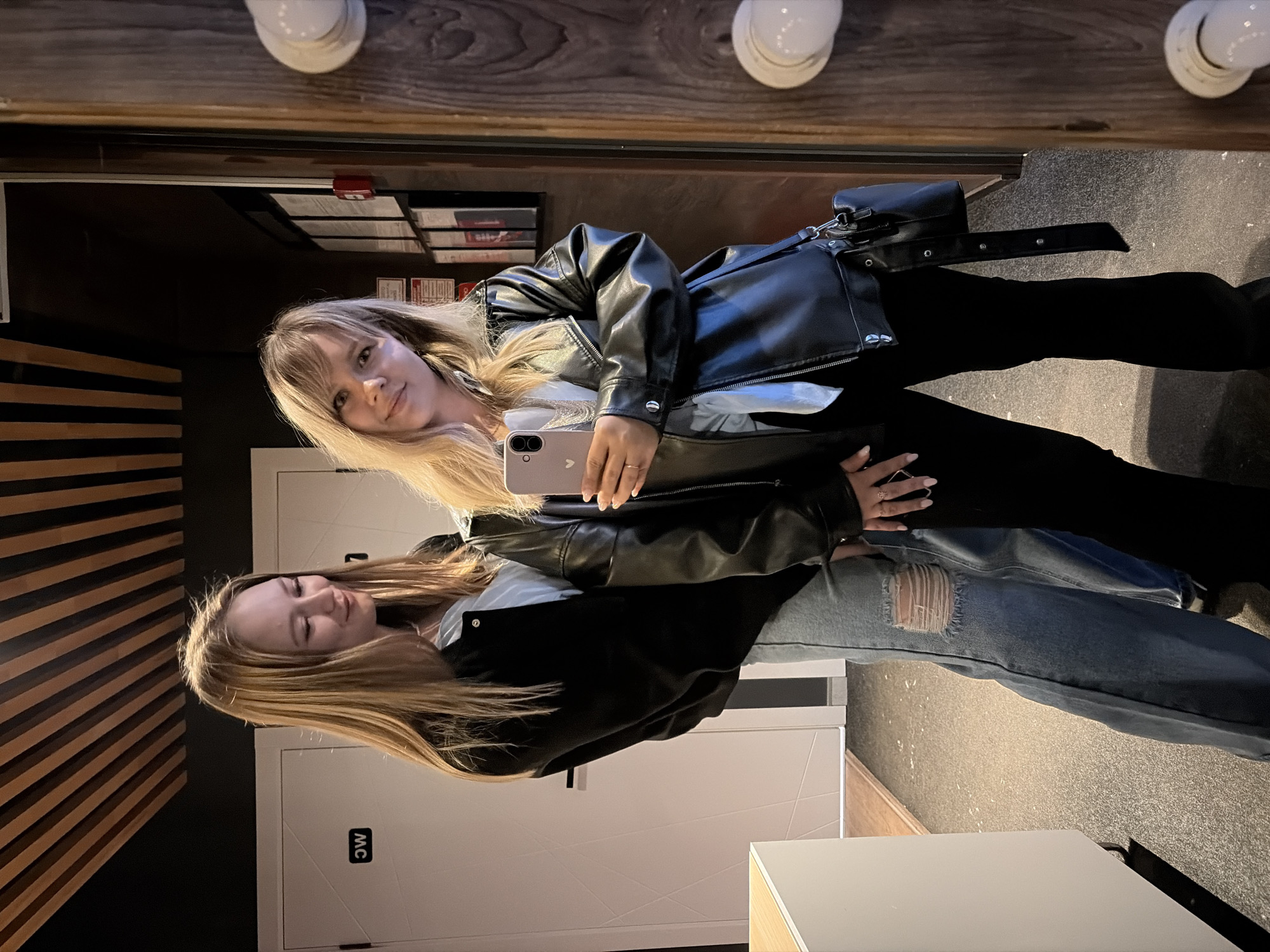 Two young women are depicted in an indoor setting, identified as the city of Chaikovsky, Russia, posing for a mirror selfie.
The woman on the left, with long, straight, light-brown hair, is wearing a black bomber jacket over a light blue shirt and distressed light blue jeans with rips at the knees. She stands slightly behind the other woman, looking towards her with a subtle smile.
The woman on the right, with blonde hair and bangs, is holding a light pink smartphone with a heart sticker on its back in her left hand, taking the photo. She is wearing a black faux leather jacket over a light-colored shirt, featuring a visible silver necklace, and black flared pants. A black shoulder bag with a long strap hangs from her shoulder. A silver ring is visible on her left ring finger, and her fingernails are painted light pink. She is smiling directly into the camera/mirror.
The background features two white doors, one displaying a rotated "WC" sign, indicating a restroom. Above, a ceiling is adorned with parallel wooden slats contrasting with dark panels. To the far right, a dark wooden wall or paneling is visible, alongside a mirror frame with two spherical white lights. This wall also holds multiple framed documents or signs, some with red accents, and a red fire alarm pull station. A white counter or cabinet is present in the lower-left. The floor is dark and speckled.
