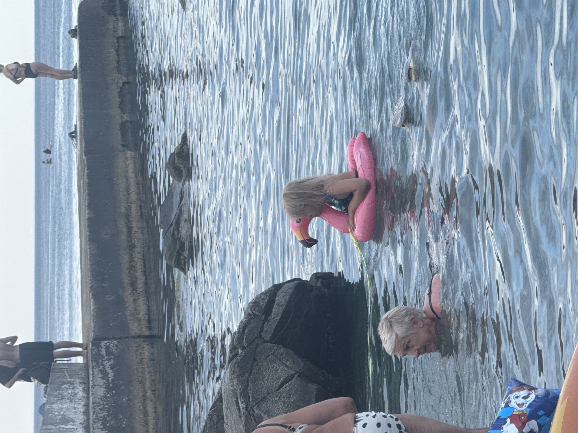 The image depicts a group of people by the sea. The scene appears to be a rocky coastline with a concrete structure, possibly a pier or seawall. The water is calm and reflects the sky, suggesting fair weather.
In the foreground, a person is lying down, partially visible, wearing a polka-dot bikini. Next to them, a child is playing with a pink flamingo inflatable. Further out in the water, an adult with short grey hair is swimming. In the background, another person is standing on the concrete structure, and a few other figures are visible further down.
The time of day appears to be late afternoon or early evening, as indicated by the soft lighting and the slight glow on the water. The overall mood is relaxed and leisurely, with people enjoying a day at the seaside. There are no visible text elements in the image. The location is identified as Partenit, Ukraine.