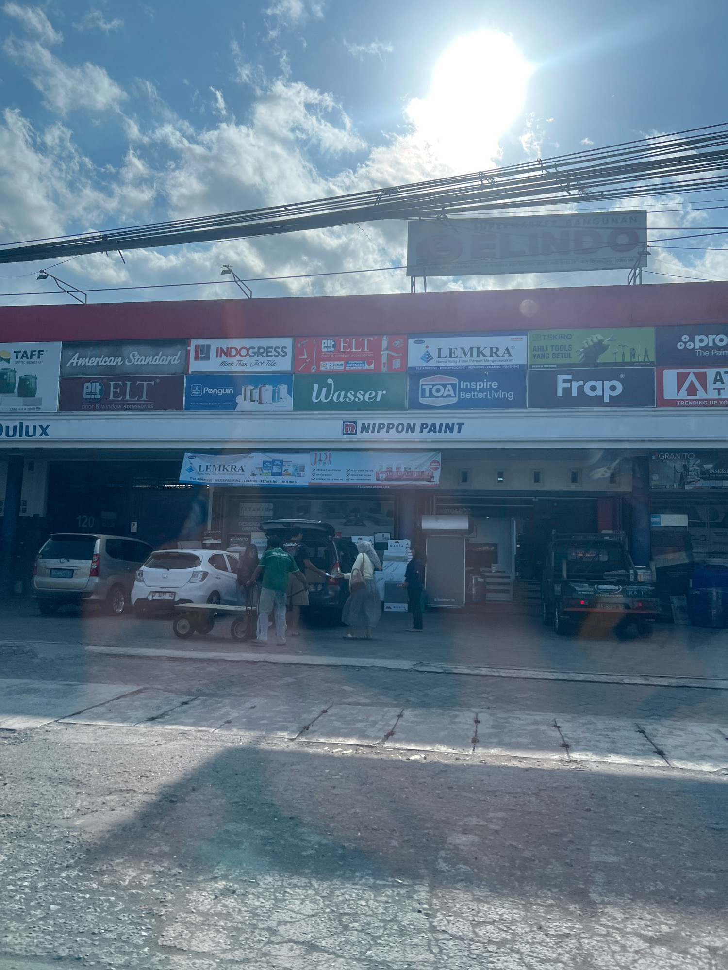 In the city of Payaman, Indonesia, a building displays multiple commercial signs. A large overhead sign reads "SUPER MARKET BANGUNAN ELINDO". Below this, a red awning supports a row of smaller signs, advertising brands such as TAFF, American Standard, INDOGRESS, ELT, Penguin, Wasser, TOA Inspire, LEMKRA, TEKIRO, Frp, and PRO.
The building's storefront is visible, with several vehicles parked outside: a silver SUV, a white hatchback, and a black pickup truck. A group of individuals are interacting near the white car and a cart, suggesting a commercial transaction or loading/unloading activity. The environment appears to be a commercial area with a road in the foreground, showing cracked pavement and a sidewalk. The sky is bright with scattered clouds and a strong sun.