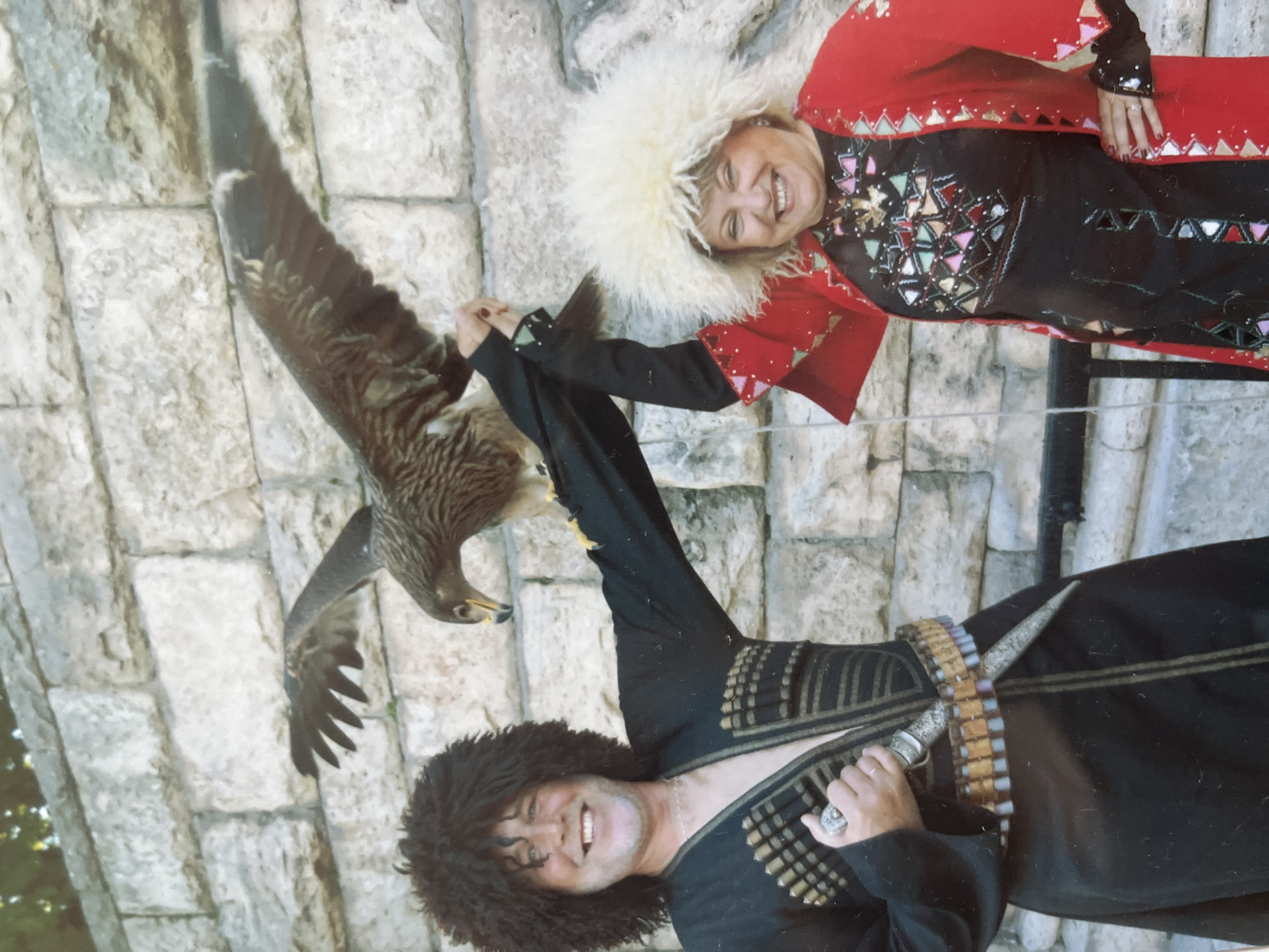 A man and a woman are posing in traditional attire in front of a stone wall. The man, on the left, has a dark, curly wig and is wearing a black outfit with decorative bullet loops and a dagger. He is smiling and holding the leg of a bird of prey perched on his arm. The woman, on the right, wears a large, fluffy white hat and a red jacket over a black dress adorned with colorful geometric patterns. She is also smiling and has her arm raised. The bird of prey appears to be a hawk or eagle. The background is a rough, light-colored stone wall. The image is a photograph, likely taken outdoors in a public space, possibly during a cultural event or festival in Vladimirovka, Russia.