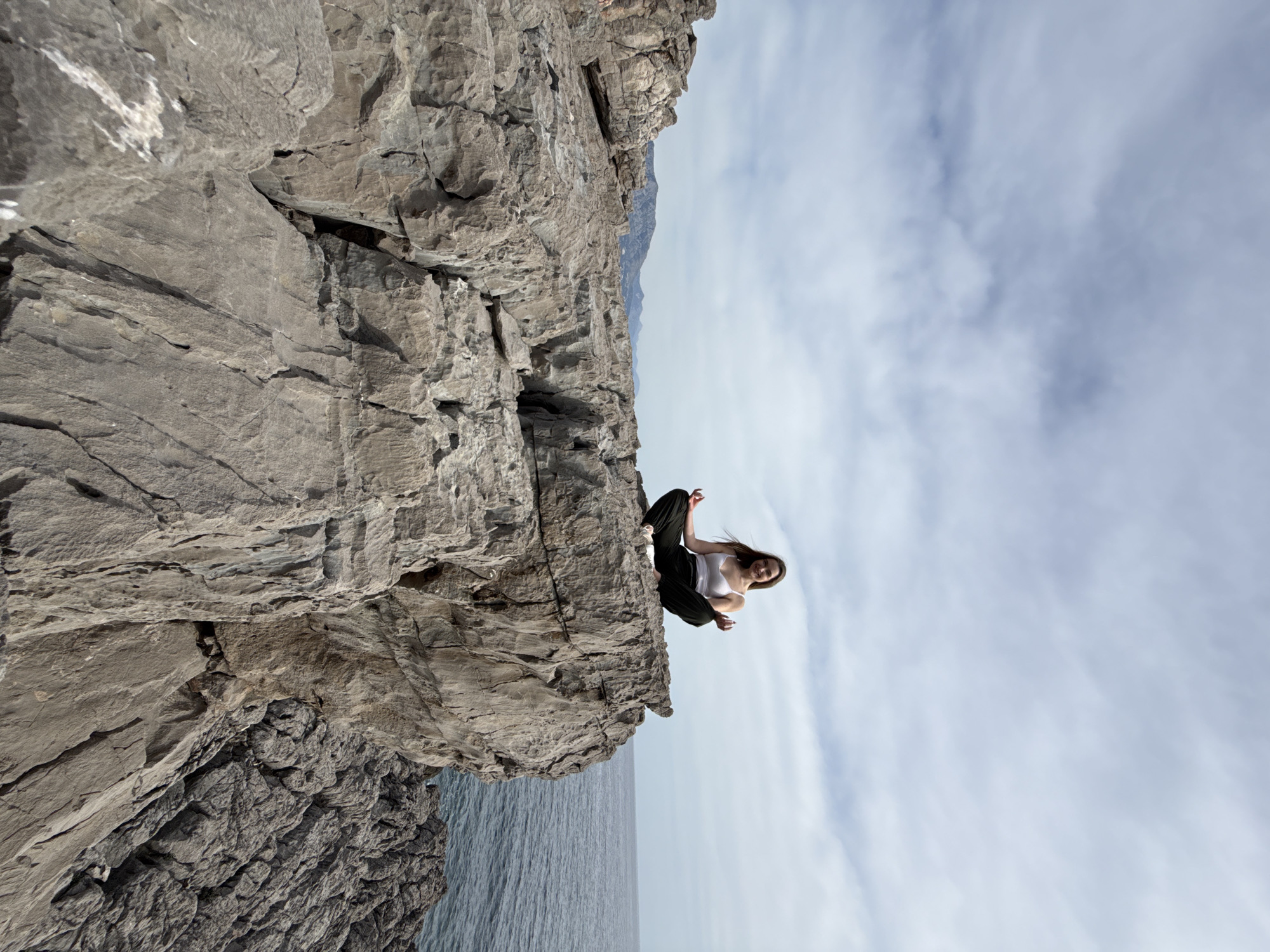 The image depicts a woman sitting in a lotus position on a rocky cliff face. She is wearing a white tank top and black pants. The cliff is gray and textured, with the sea visible below and a cloudy sky in the background. The location is Boreti, Montenegro.