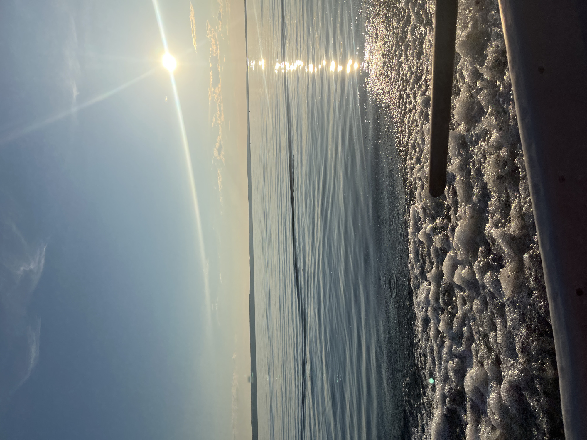 This image depicts a serene scene of a body of water, likely a lake or river, with a clear sky. The sun is prominently featured, casting a bright glare and reflecting on the water's surface, creating a shimmering effect. The water appears calm, with gentle ripples indicating a possible breeze. On the right side of the image, the wake of a boat is visible, characterized by foamy, disturbed water and two metallic poles, suggesting the perspective is from on board a vessel. The sky is a soft blue with a few wispy clouds. The time of day appears to be either early morning or late afternoon due to the low angle of the sun and the warm, golden light. The overall mood is peaceful and tranquil, evoking a sense of calm and relaxation. The location is identified as Siukterka, Russia, which could be a specific village, town, or natural area within Russia. There is no visible text in the image.