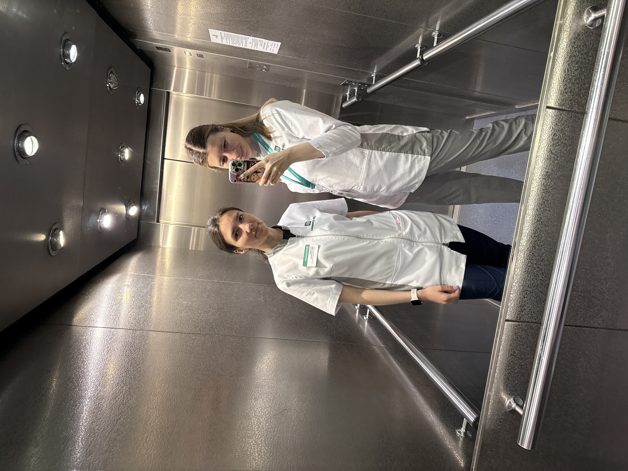 Two young women are captured in a selfie taken inside a modern elevator. The elevator's interior walls feature a combination of brushed metal panels and textured dark grey/black surfaces, with circular recessed lights illuminating the space from the ceiling. A metal handrail is visible along one wall, and the floor also appears to be textured metal.
The woman on the right, positioned slightly forward, is holding a smartphone with a decorative, cartoon-themed case to take the picture. She is smiling and looking at the phone's screen. She wears a white professional jacket or lab coat over grey trousers, and a green lanyard is visible around her neck.
The woman on the left stands slightly behind her, also smiling and looking towards the camera (the phone). She is wearing a similar white jacket or lab coat over dark blue jeans and has a white smartwatch on her left wrist. A visible ID badge on her jacket displays the text "ЦУП ИГРА" (TSUP IGRA) and a green, stylized logo. Both women appear relaxed and content.
On the elevator wall behind them, a white notice with text in Russian is partially visible, likely an information or instruction sign typical for elevators. The scene depicts a casual moment, possibly between colleagues or friends, within an elevator setting in Moscow, Russia.