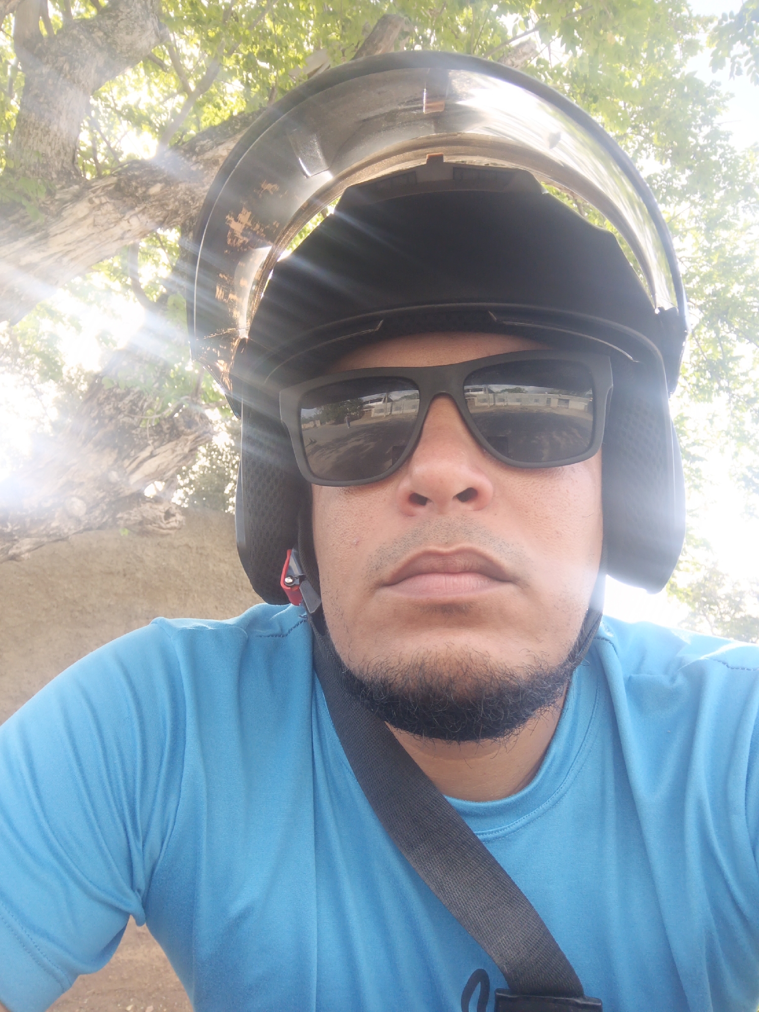 A close-up, waist-up, outdoor, selfie-style photograph shows a man wearing a black motorcycle helmet and dark sunglasses. He has a light beard and is wearing a bright blue t-shirt. A black strap runs diagonally across his chest. The background consists of a large tree with green leaves and sunlight filtering through the branches, suggesting a bright, sunny day. The terrain behind him appears to be a rough, sandy or dirt surface. In the reflection of his sunglasses, buildings are visible, possibly indicating an urban or suburban setting. The image was taken in Juan Griego, Venezuela.