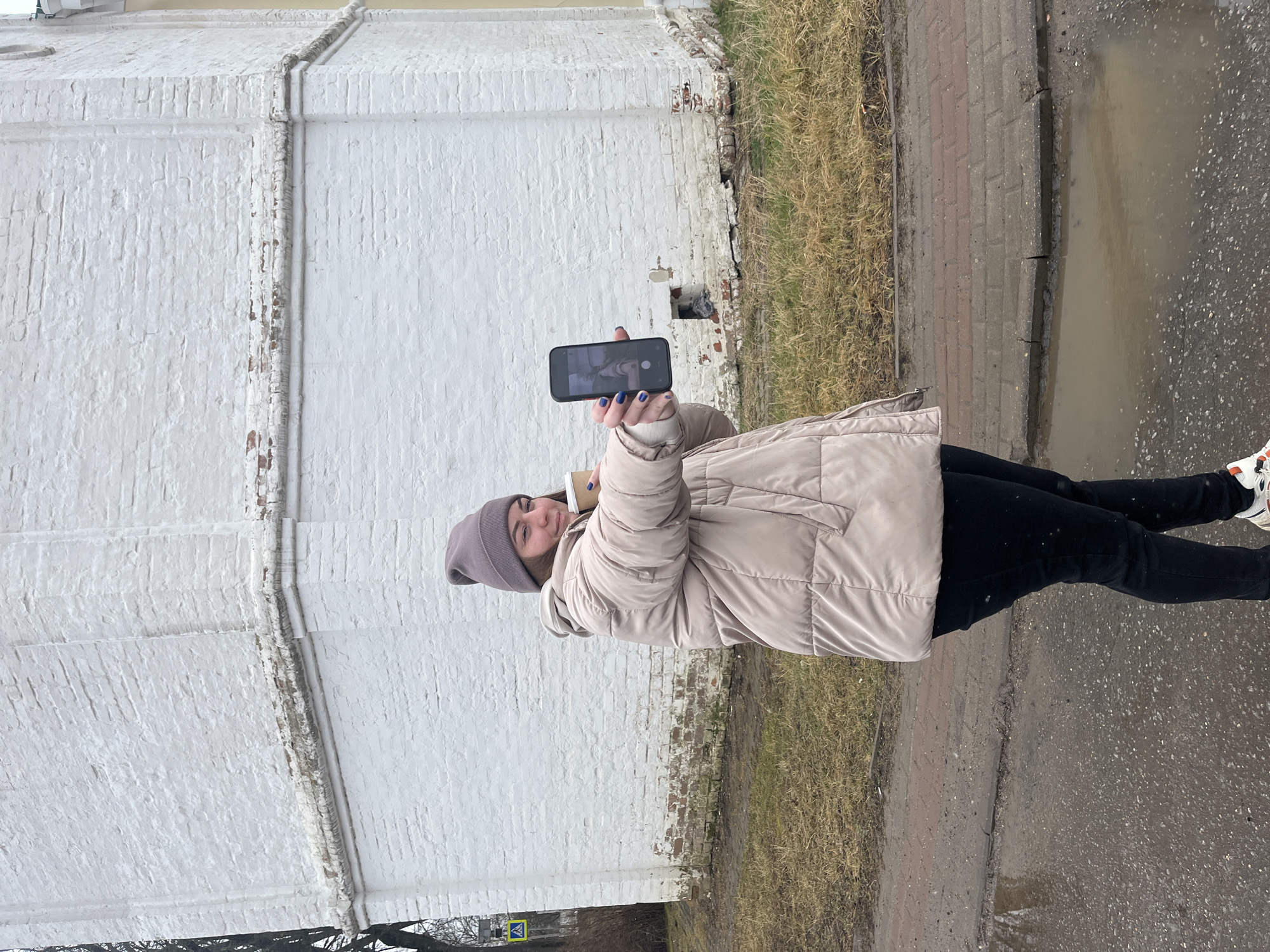 A woman stands on a paved sidewalk in Lugovoy, Russia. She wears a beige puffer jacket, black leggings, a gray beanie, and white sneakers. She holds a smartphone in her left hand, apparently taking a photo of herself. In her right hand, she holds a coffee cup. Behind her is a whitewashed brick wall. To the right, adjacent to the sidewalk, there is a patch of dry grass and a large puddle reflecting the gray sky. The pavement is wet. At the bottom left of the image, a pedestrian crossing sign is visible.
