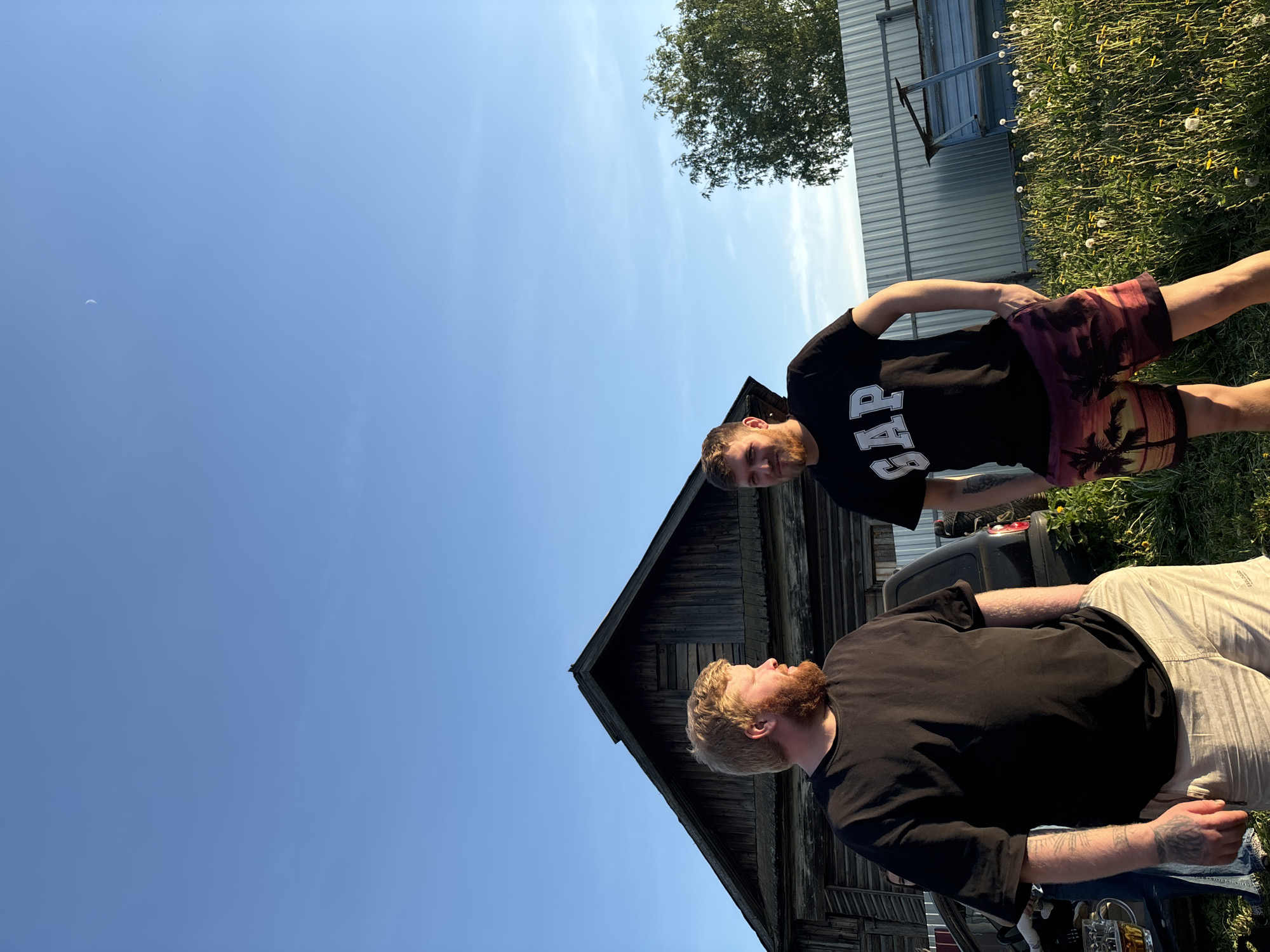 Two Caucasian men are standing outside an old, wooden building on a sunny day. One man wears a black "GAP" t-shirt and patterned shorts. The other man is wearing a black t-shirt and light-colored pants. A silver building and trees are visible in the background. The location is most likely rural, but exact geographical data cannot be definitively confirmed.