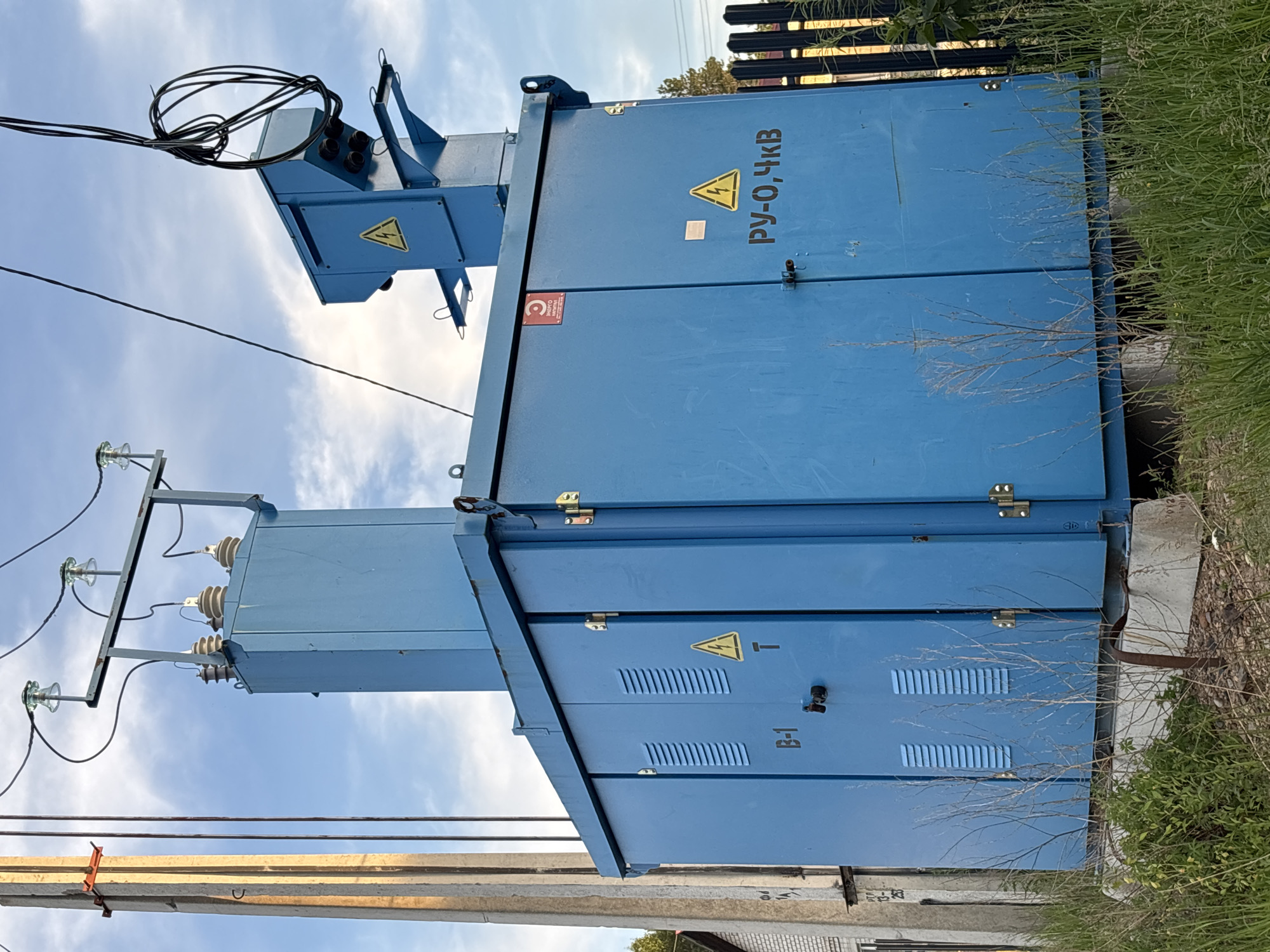 The image shows a large blue electrical transformer box situated outdoors, likely in a utility area. The transformer box is tall and rectangular, with several doors and vents on its sides. A smaller, tiered blue structure with a junction box containing coiled cables is mounted on top of the main box. Electrical insulators and wires are visible connecting to the top of the transformer and extending towards a concrete utility pole on the left side of the frame. A yellow triangular warning symbol with a lightning bolt is visible on the front of the transformer. Cyrillic text, including "РУ-О,ЧКВ", is also present on the box. The transformer is surrounded by tall grass and dry weeds, with a black metal fence visible on the right. The sky above is partly cloudy. There are no people visible in the image. The setting appears to be in a rural or semi-urban area, possibly in the city of Chigiri, Russia, given the presence of Cyrillic text and the style of the utility equipment.