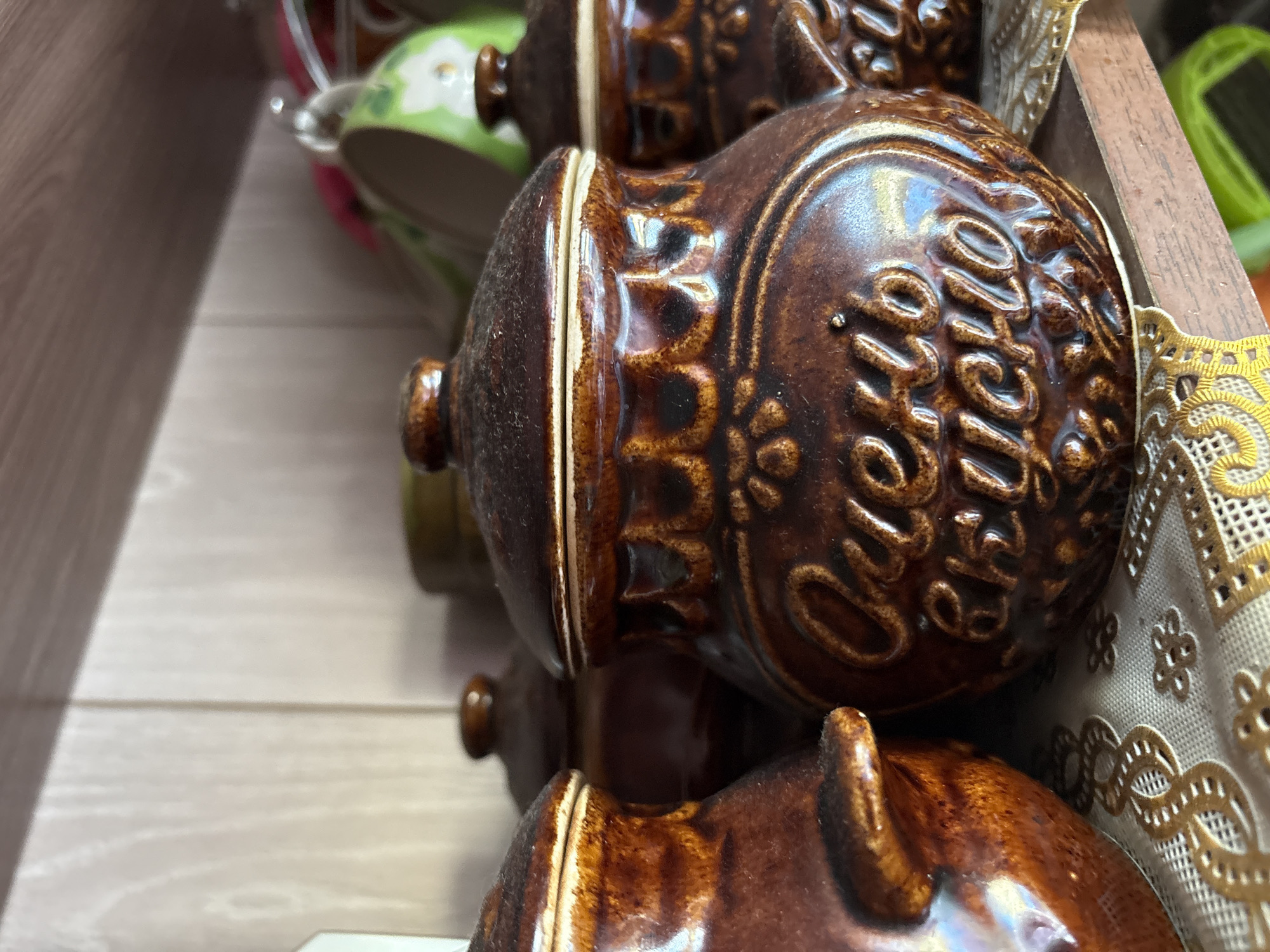 A close-up, eye-level shot captures three brown ceramic jars with lids on a wooden shelf. The jar in the foreground, slightly off-center to the right, is ornately decorated with embossed Cyrillic lettering that reads "Очень вкусно" (Very Tasty). It has a rounded body and a domed lid with a small knob. To its left, another similar jar is partially visible, and further to the left, a third jar's side is in view. Behind these jars, other objects are blurred, including a green ceramic mug with a floral pattern and the suggestion of more jars or containers. The shelf is covered with a textured white tablecloth adorned with a gold decorative trim. The background consists of a light-colored, wood-paneled wall. The lighting is soft, creating gentle highlights on the glossy ceramic surfaces. The scene appears to be an indoor setting, possibly a kitchen or pantry. The image is devoid of any individuals or apparent actions. This scene is likely located within the city of Vetluzhsky, Russia, given the context of the text on the jar.